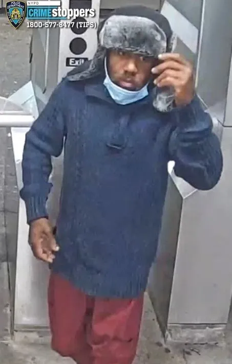 Photo of a male suspect wearing a black and gray fur-lined hat, a blue sweater, a light blue surgical mask pulled down around his chin, and red pants, standing in a subway station.