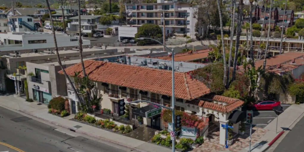 An aerial view of the 14 West Hotel, Holiday Inn Laguna Beach, a building with a red-tiled roof and black awnings, surrounded by other buildings and palm trees.