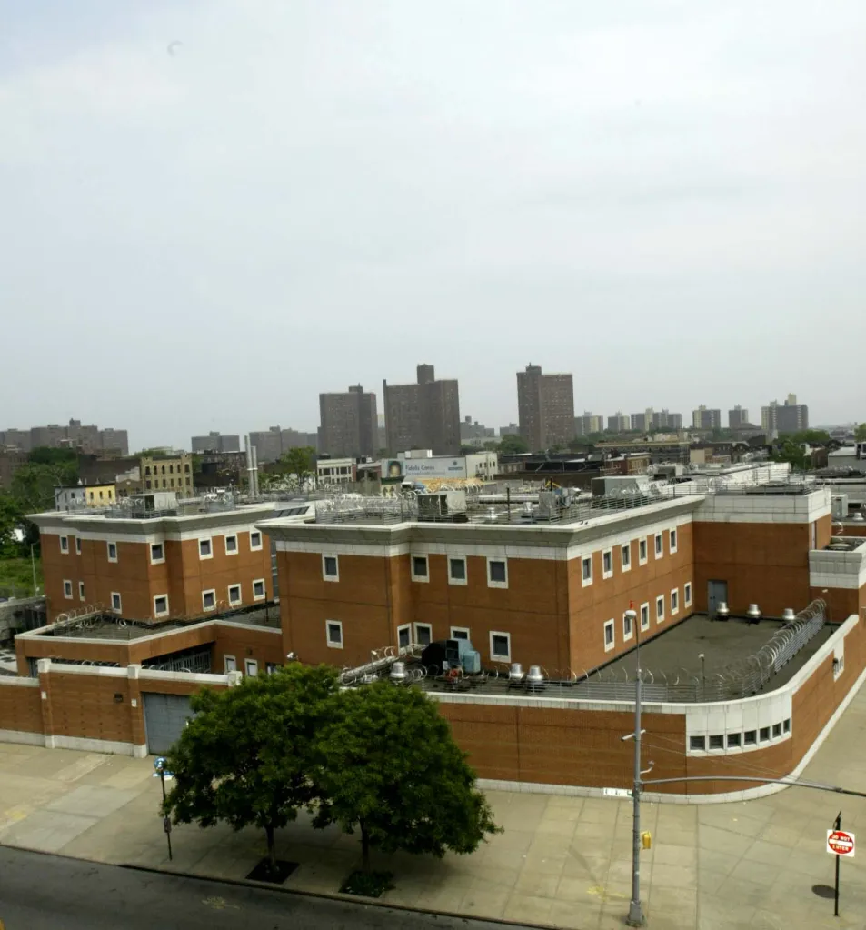 The Crossroads Juvenile Center in Brooklyn, a brown brick building with a wall topped with barbed wire, stands in front of several apartment buildings.