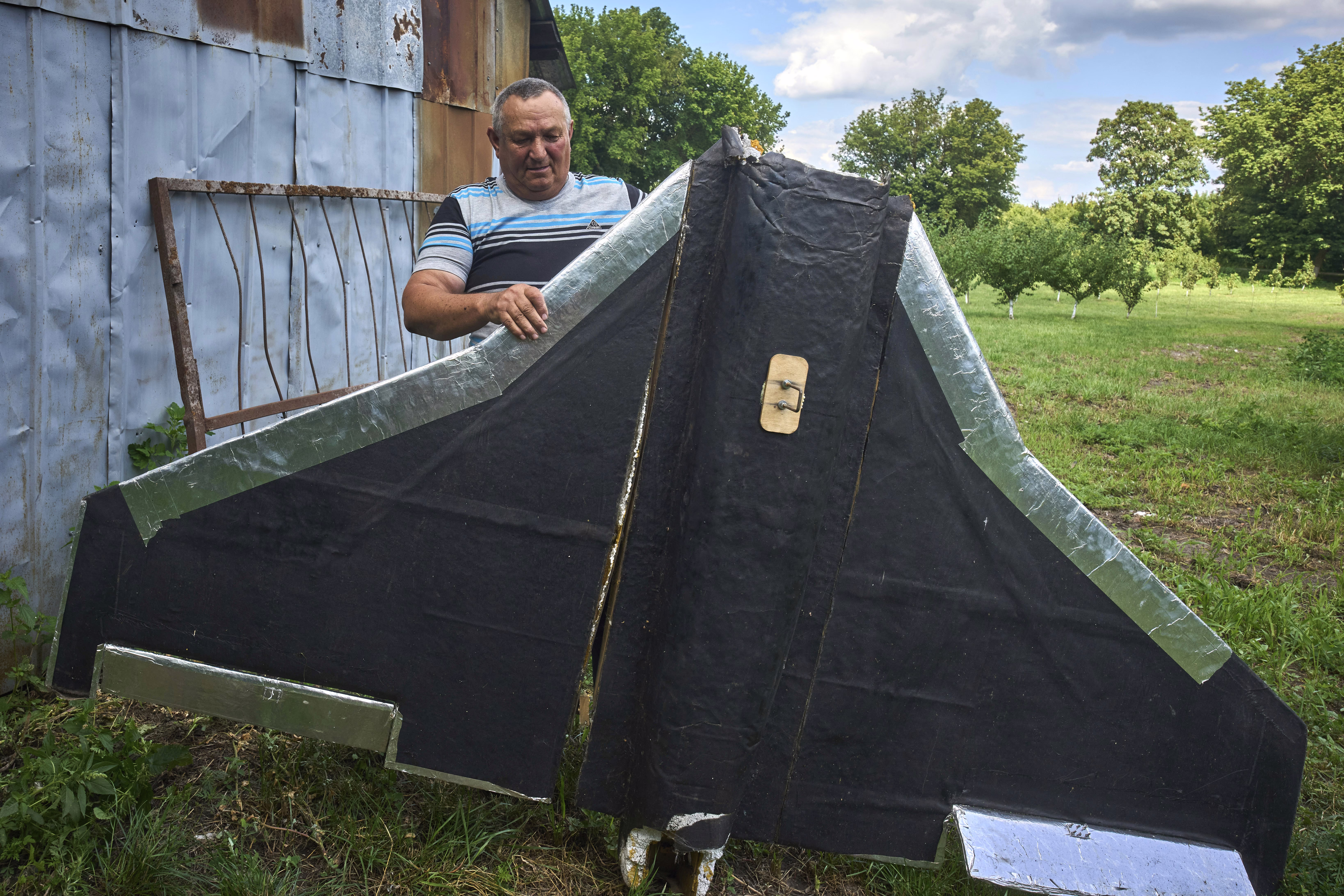 A farmer stands near a barn, holding up a large wing of a downed Russian drone.