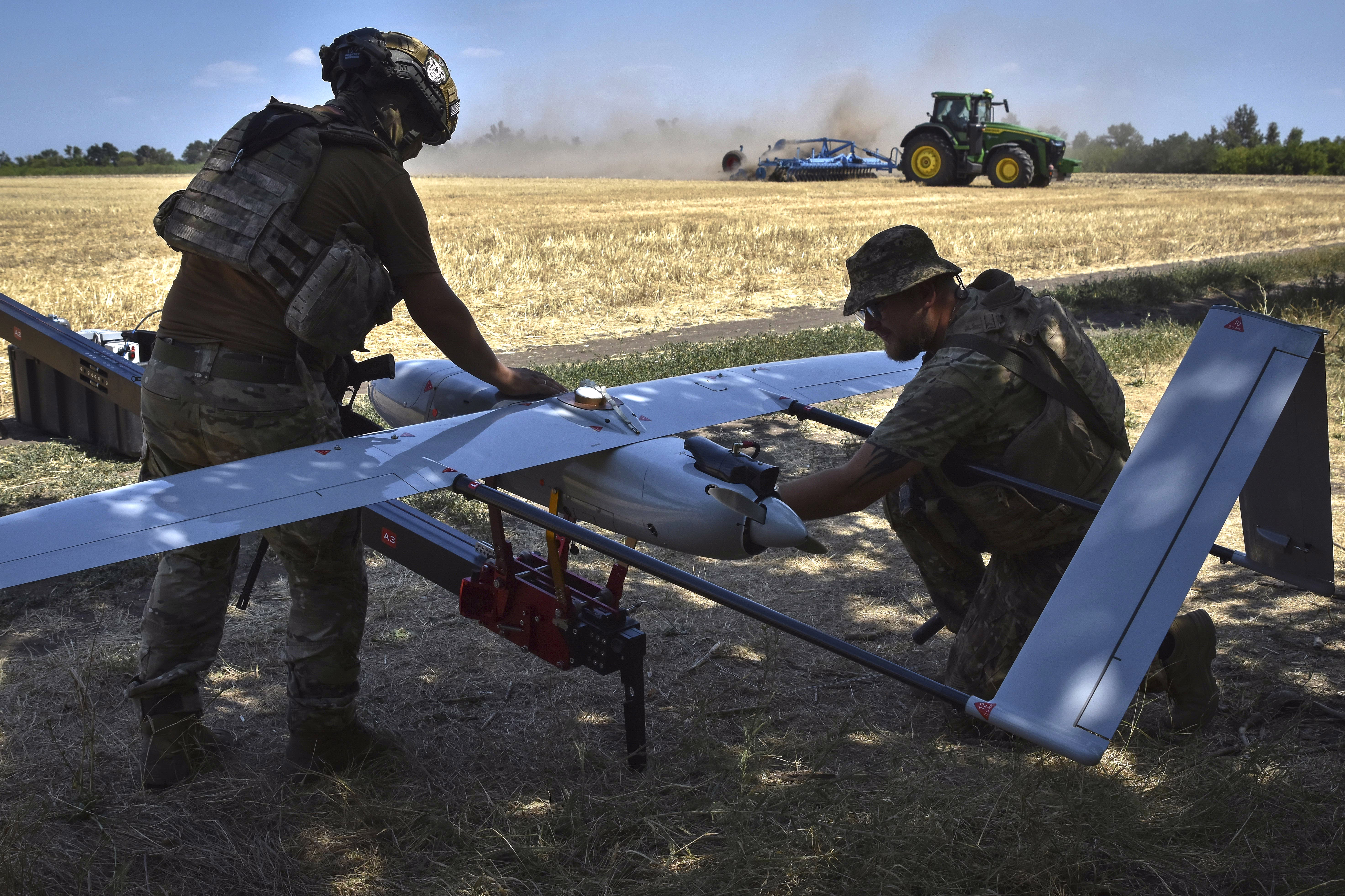 Two soldiers prepare a fixed-wing drone for a launch, beside a farm field with a tractor operating in the background.