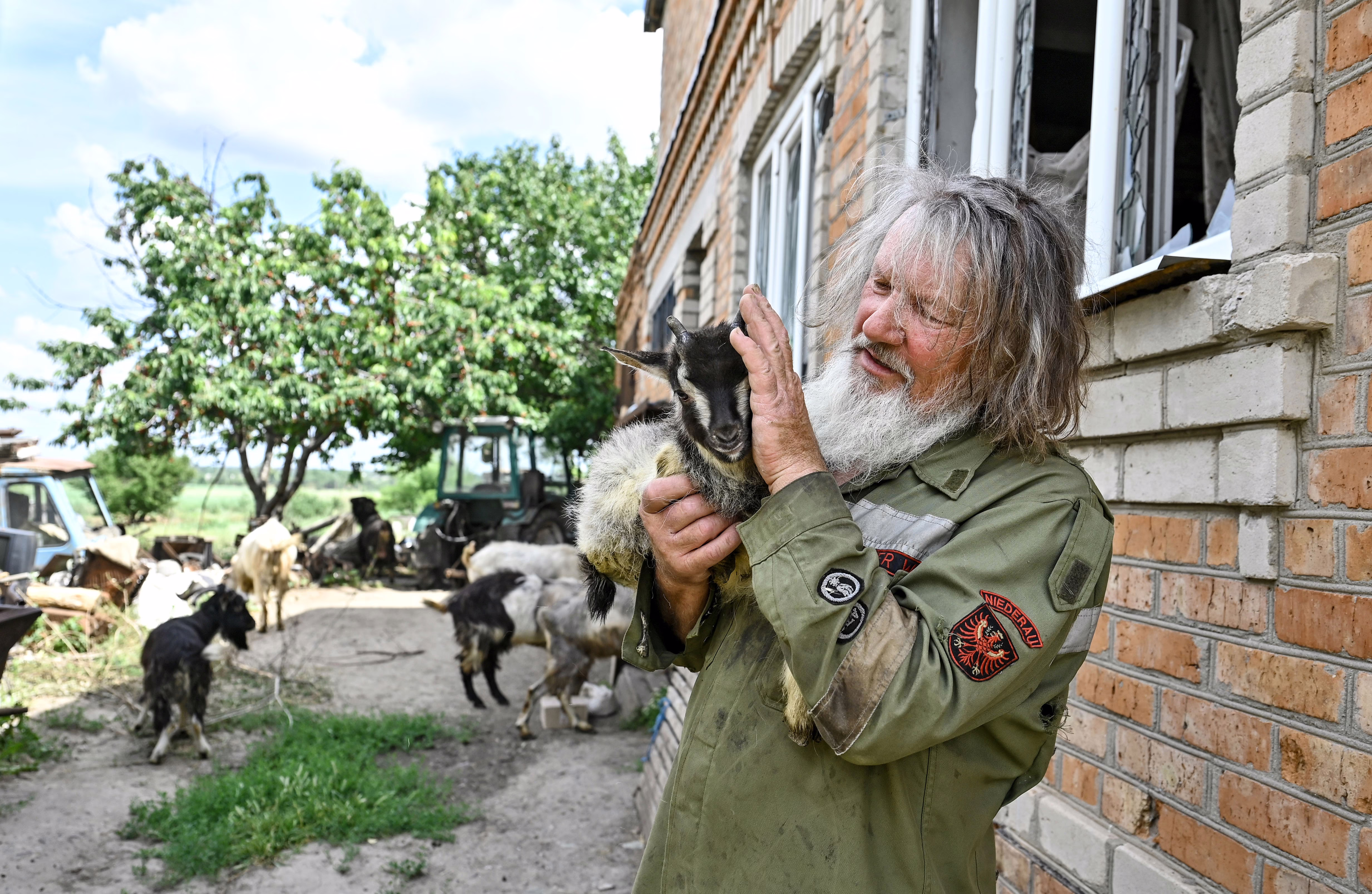 A man holds a young goat, standing near other goats beside a brick building.
