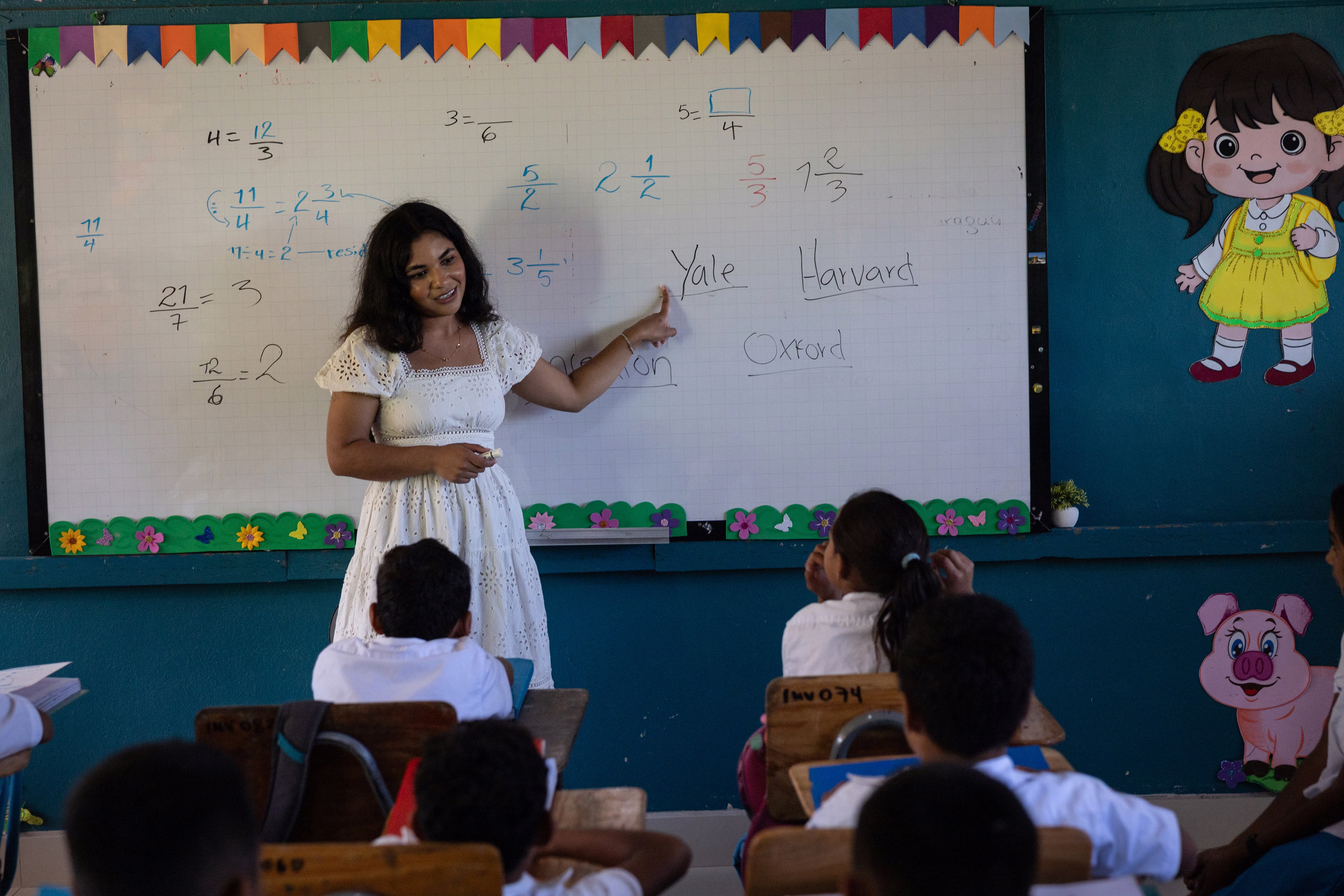 A young woman stands in front of a whiteboard, speaking to students in a classroom.