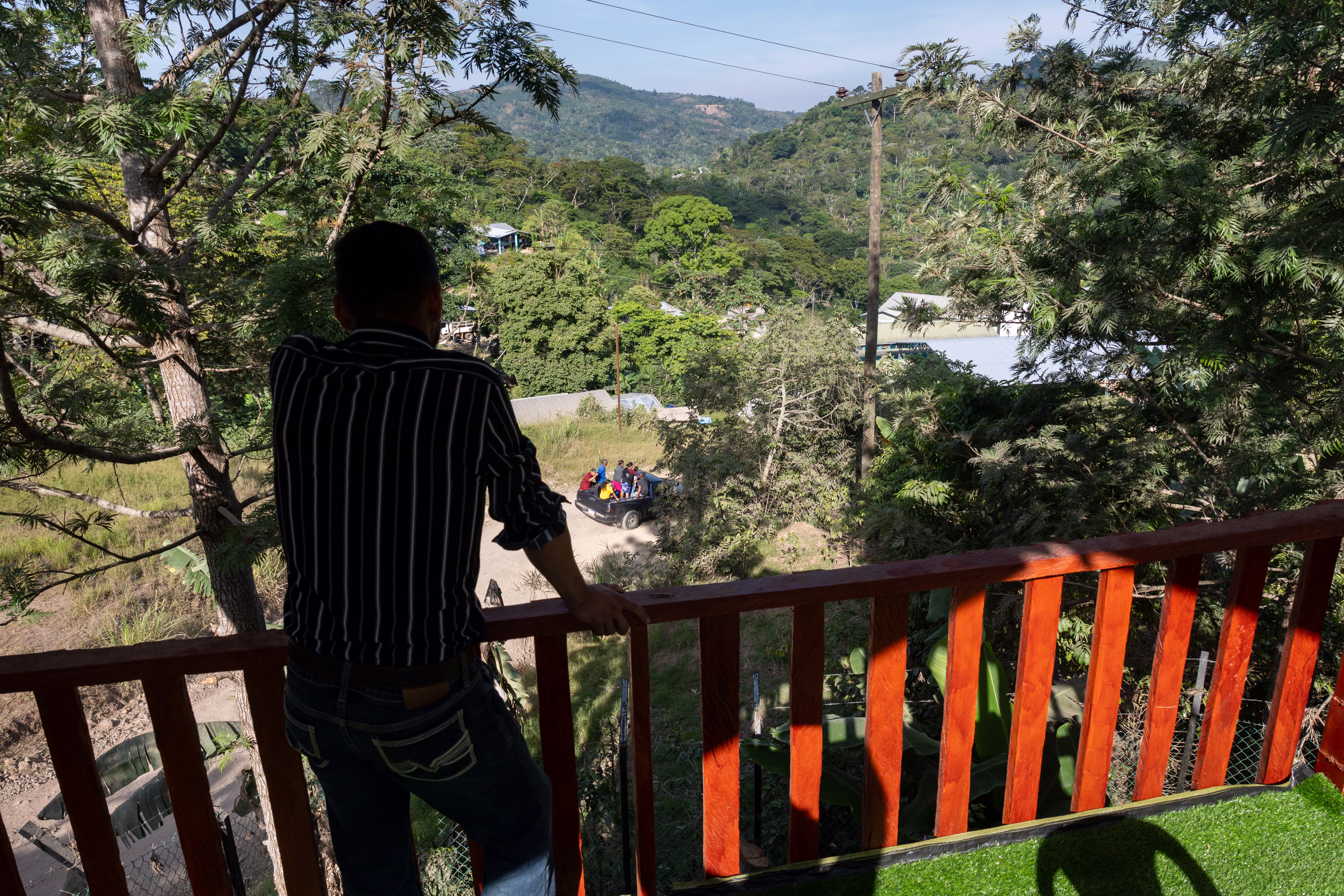 A man stands on a balcony, watching farm workers pass by below in a pickup truck.