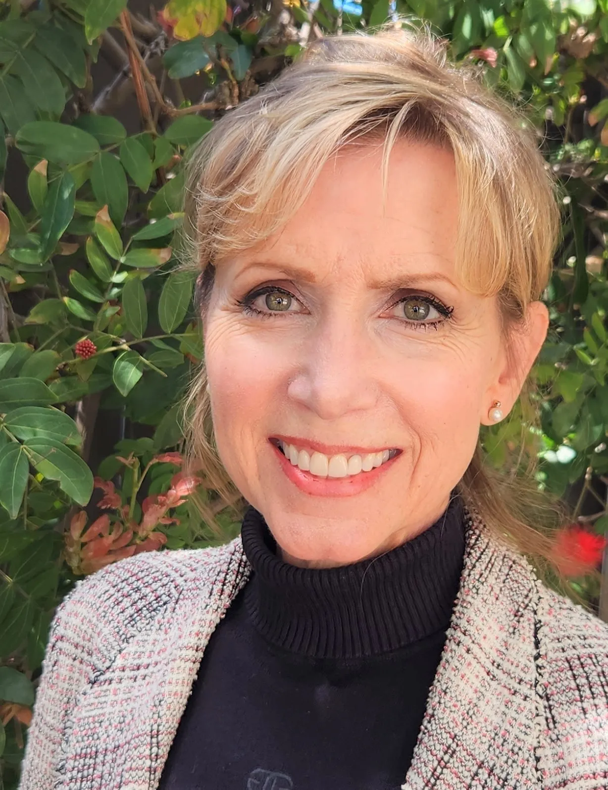 A smiling woman with blonde hair, green eyes, and a black turtleneck stands in front of green foliage.