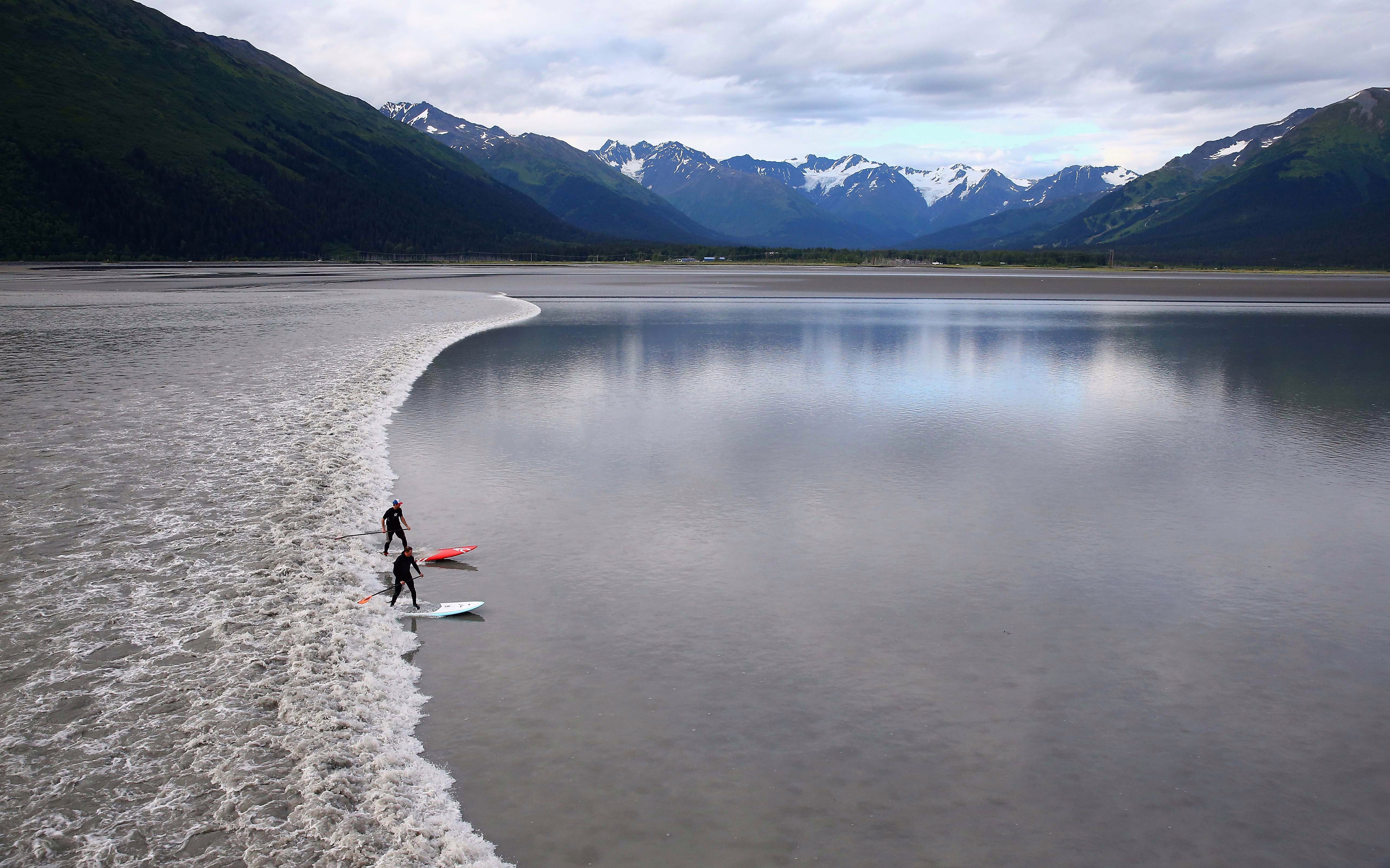 A pair of surfers catch a bore tide—a wide single wave flowing through a relatively narrow and shallow channel, seen among nearby mountains.