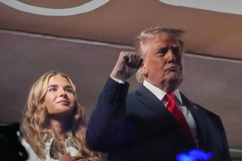 Donald Trump, in a dark suit and red tie, stands next to a blonde woman with Kai Trump, his fist raised.