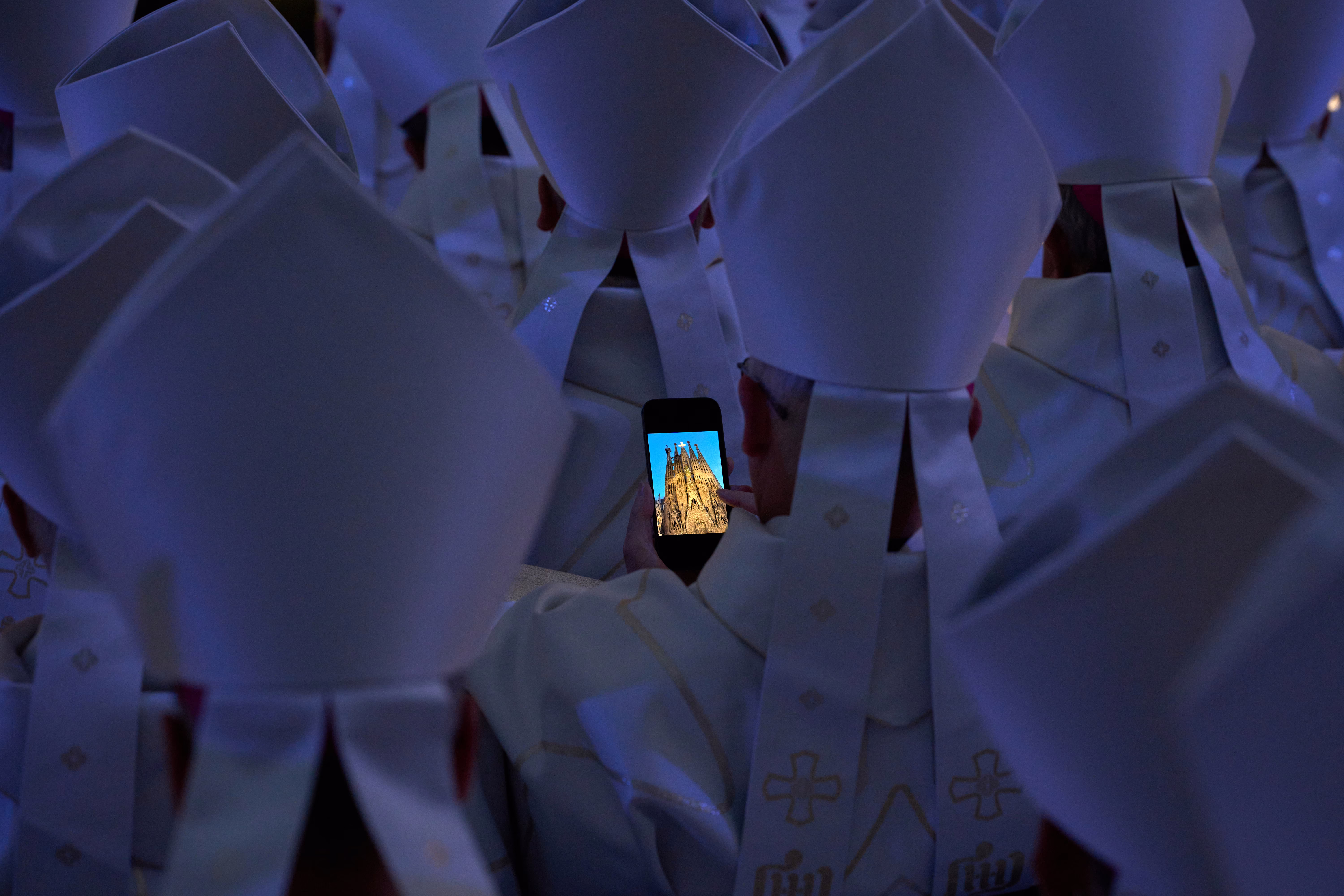 Priests in ceremonial gear gather during a ceremony, with one holding a cell phone that displays a photograph of a cathedral.