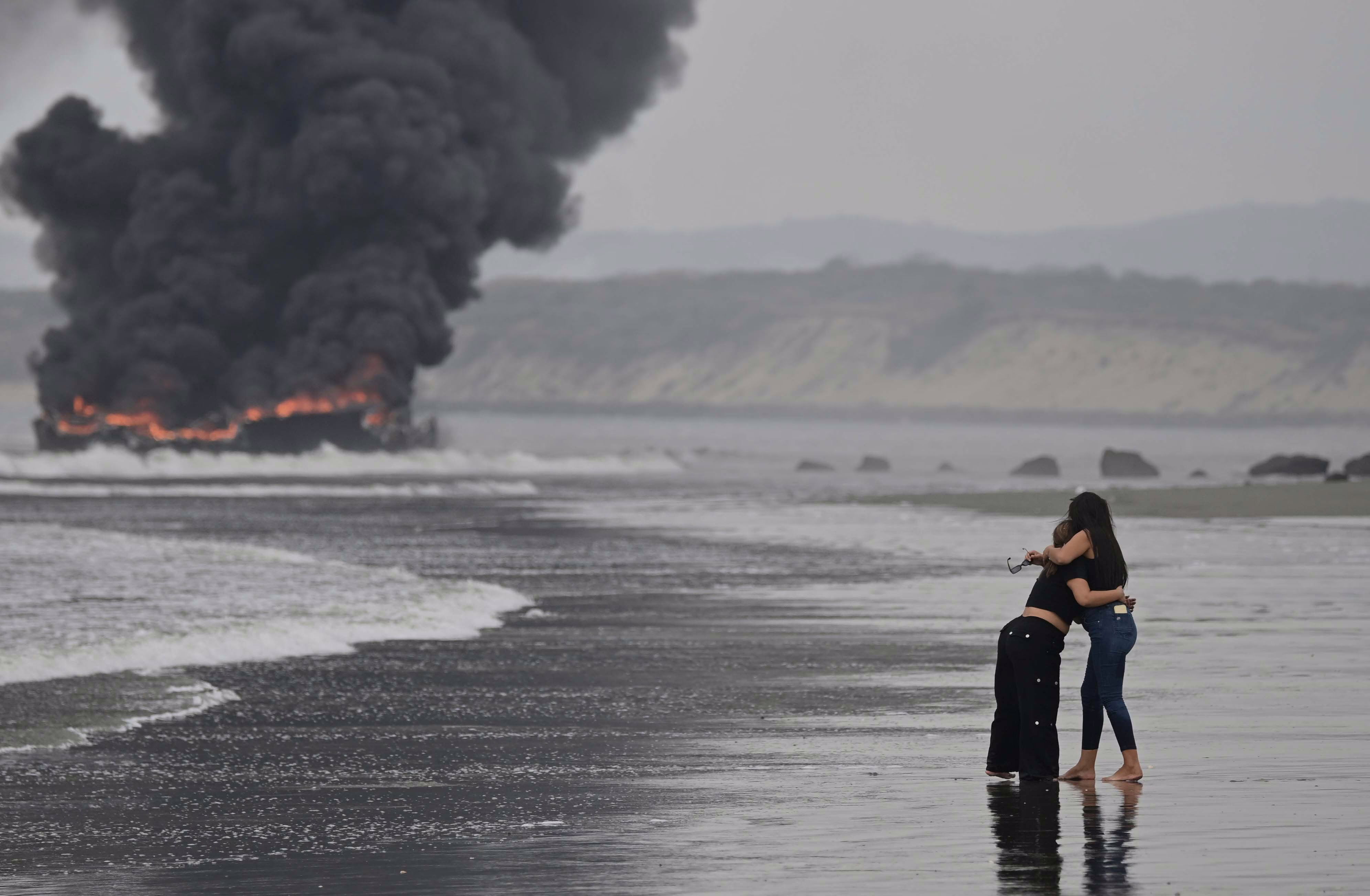 Two people embrace, standing on a beach, looking toward boats that are fully engulfed in flames in the distance.