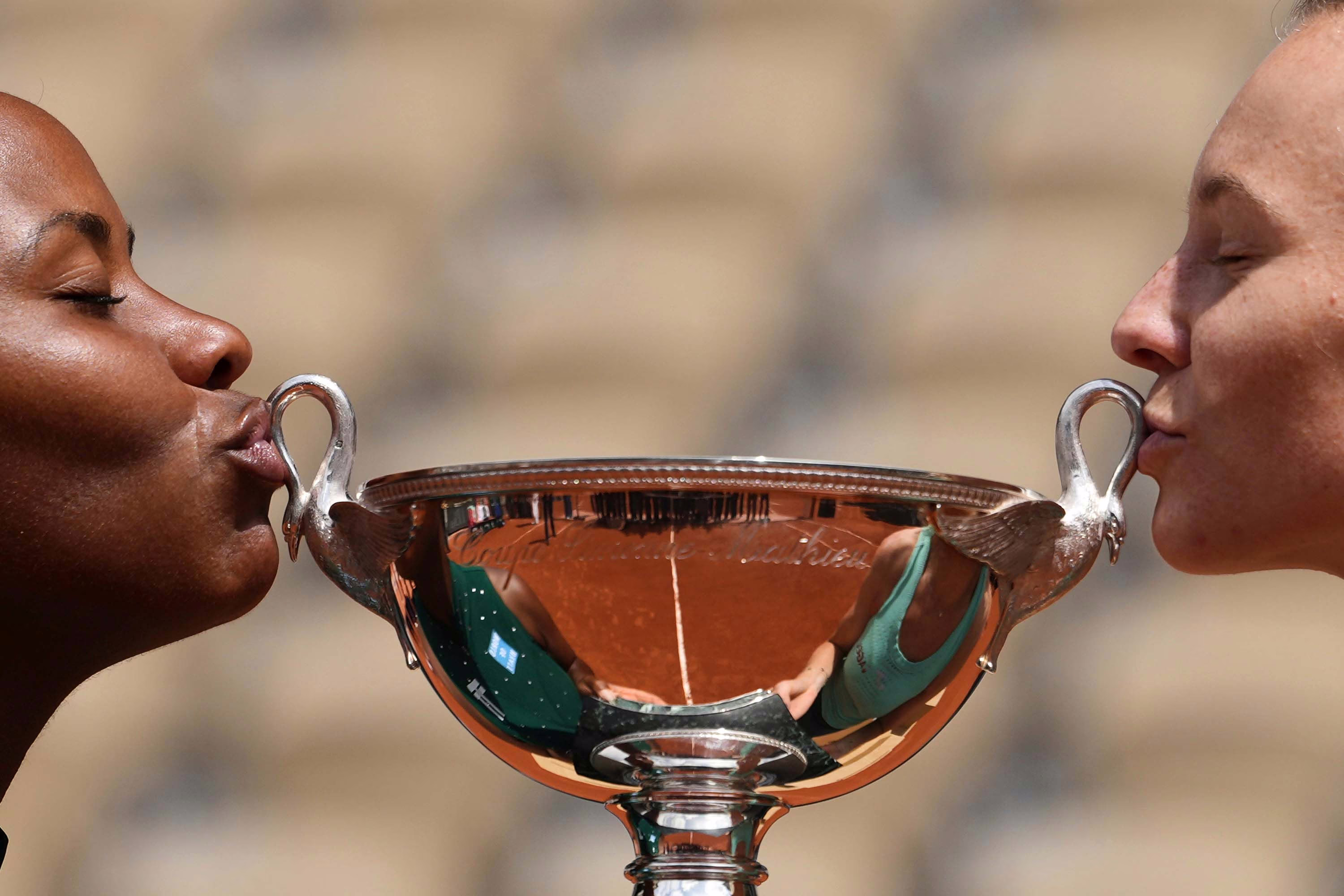 Two tennis players lean to kiss the handles of a trophy they have just won.