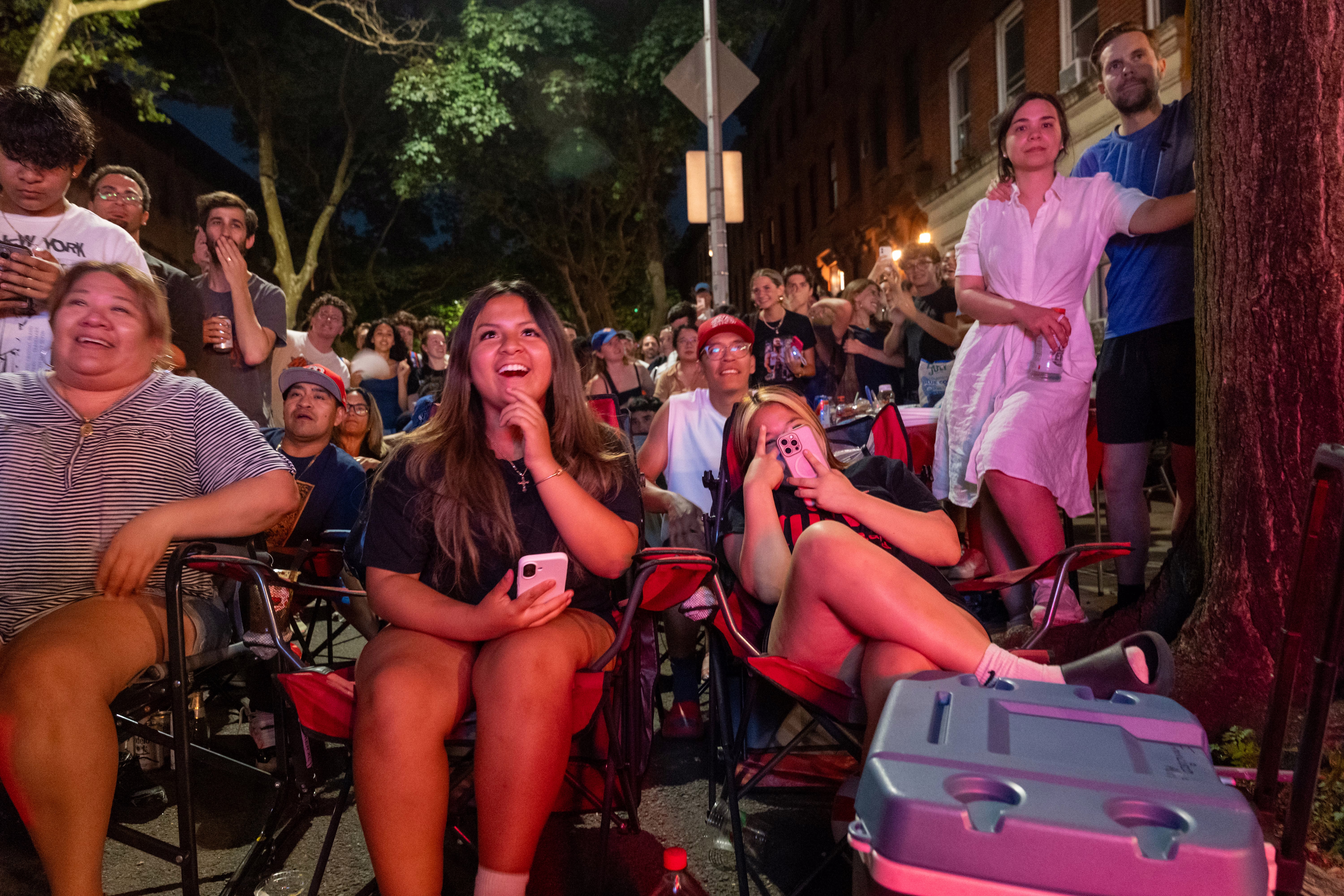 People sit and stand in a city street, watching a televised basketball game.