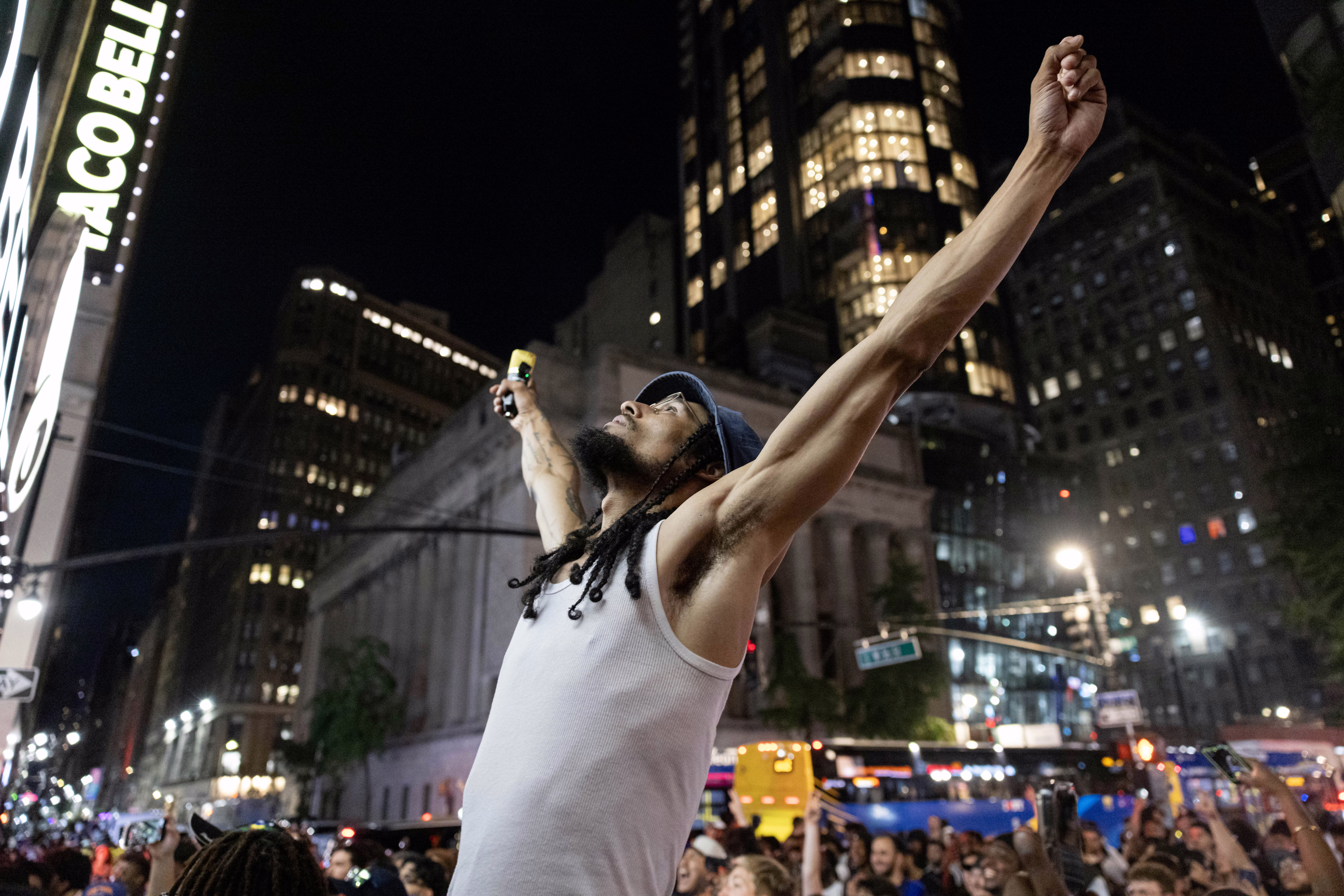 Knicks fans attend a watch party in a city street, with one fan standing at center, arms raised up.