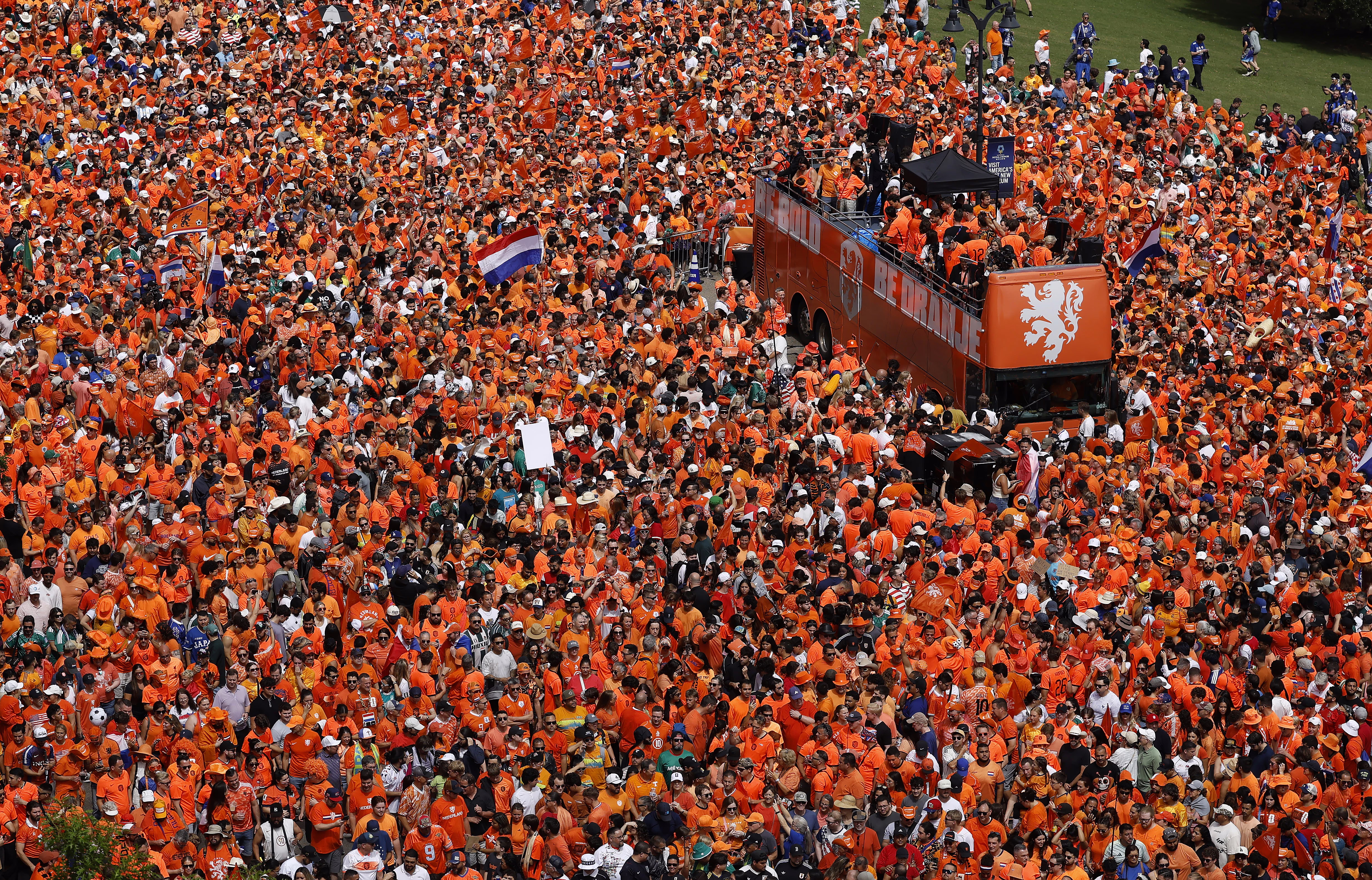 An elevated view of a large crowd of soccer fans, almost all wearing orange, gathered around a double-decker bus