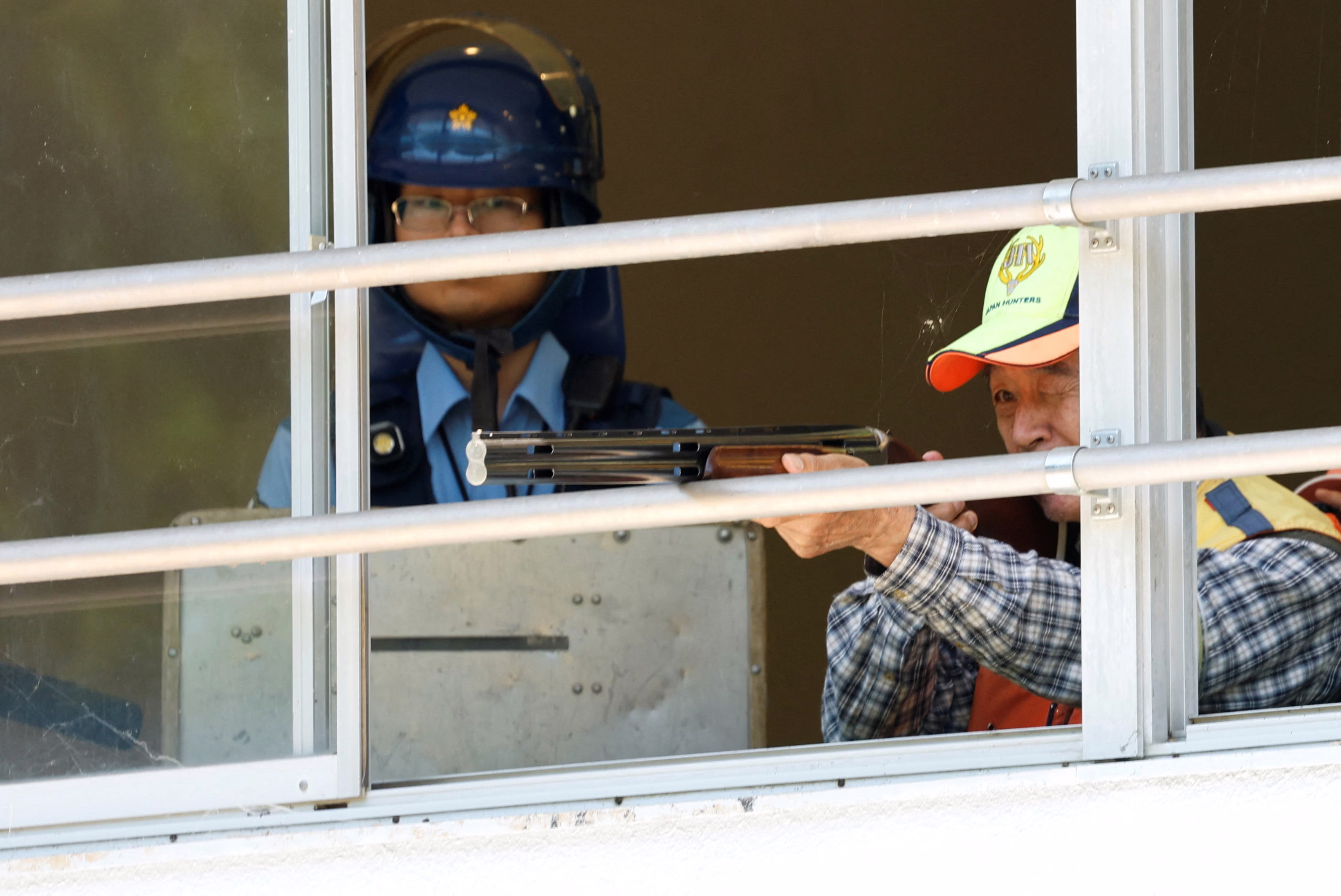 A hunter points a rifle through an open window, with a police officer watching nearby.