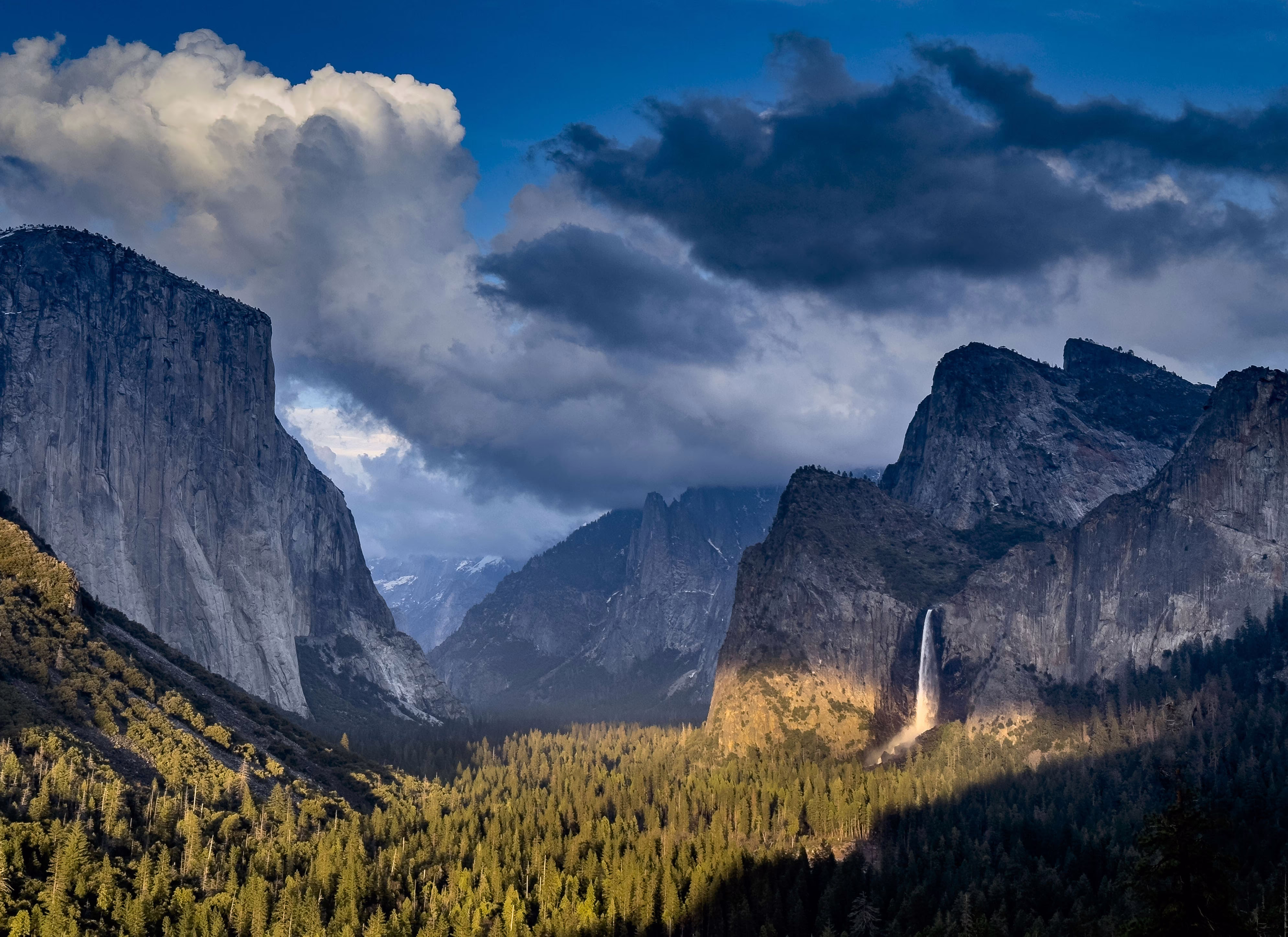 Stark and dramatic cliffs and mountains rise above a forested valley floor, with a distant high waterfall to the right of center.