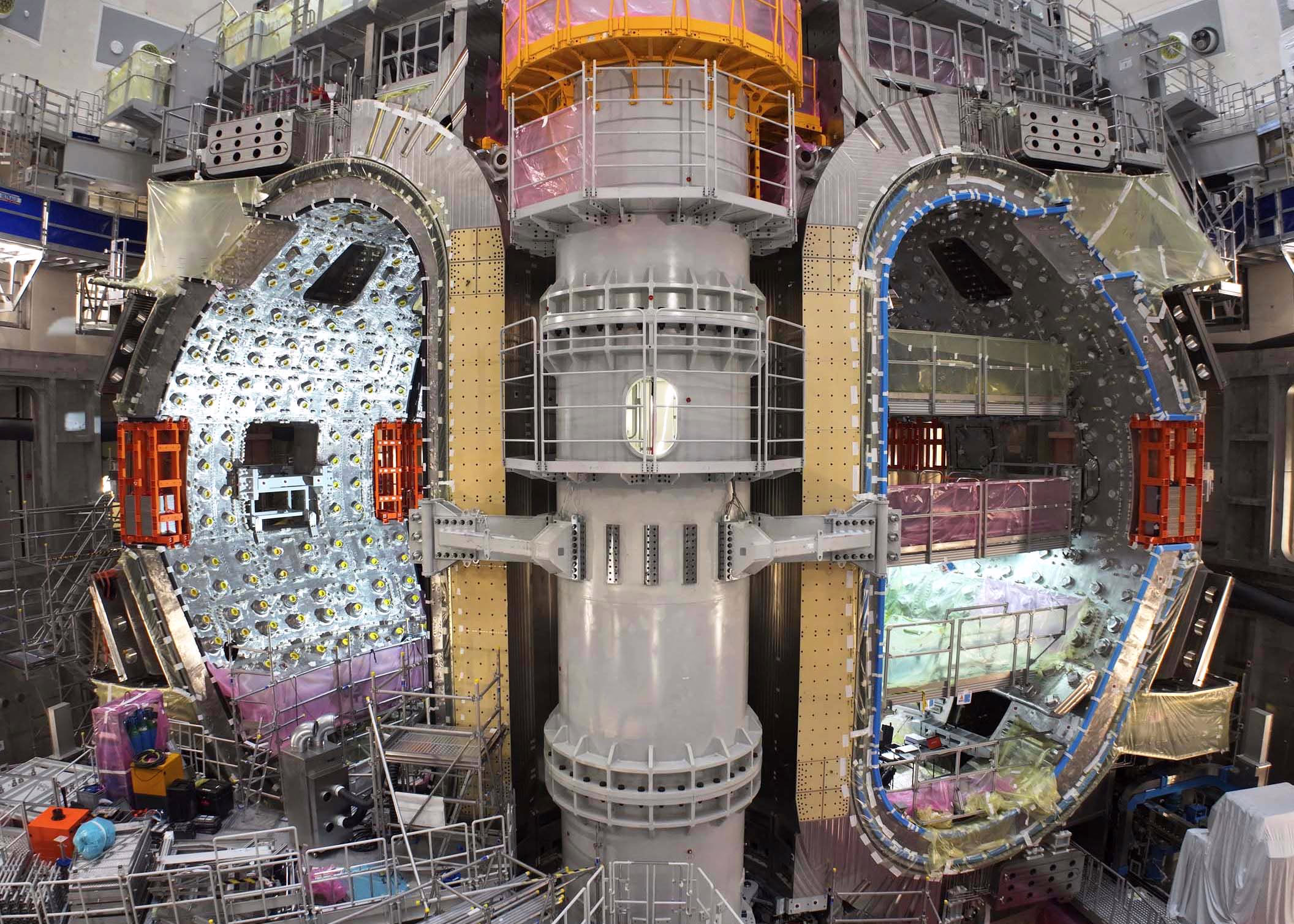 A massive fusion reactor, seen under construction. Two tall, metal wedge-shaped, or D-shaped modules sit on either side of a thick central pillar. The interior of each wedge bristles with mounting points for instruments and shielding.