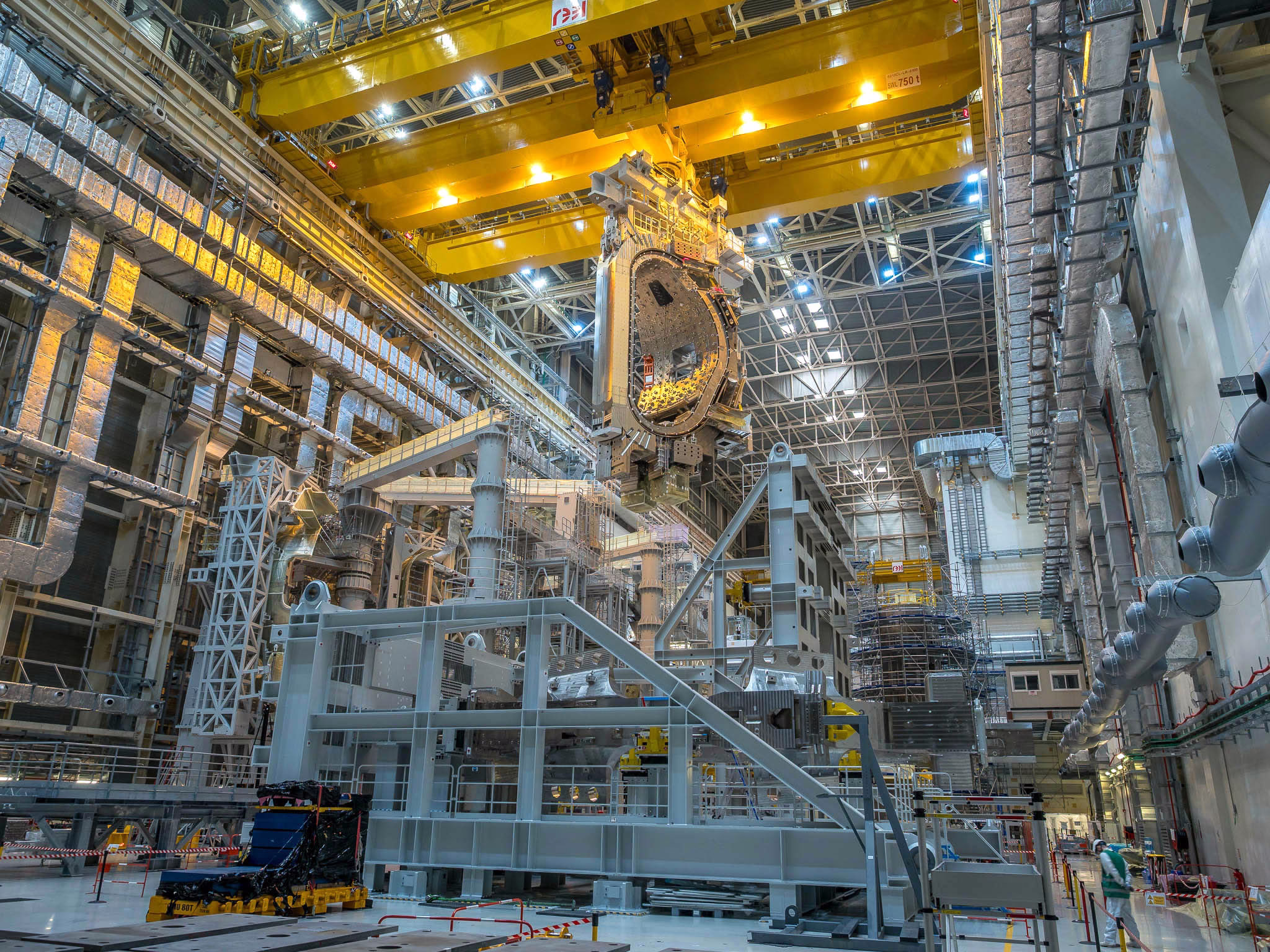A large D-shaped component of a fusion reactor is lowered into place among many supports and other partially complete components.