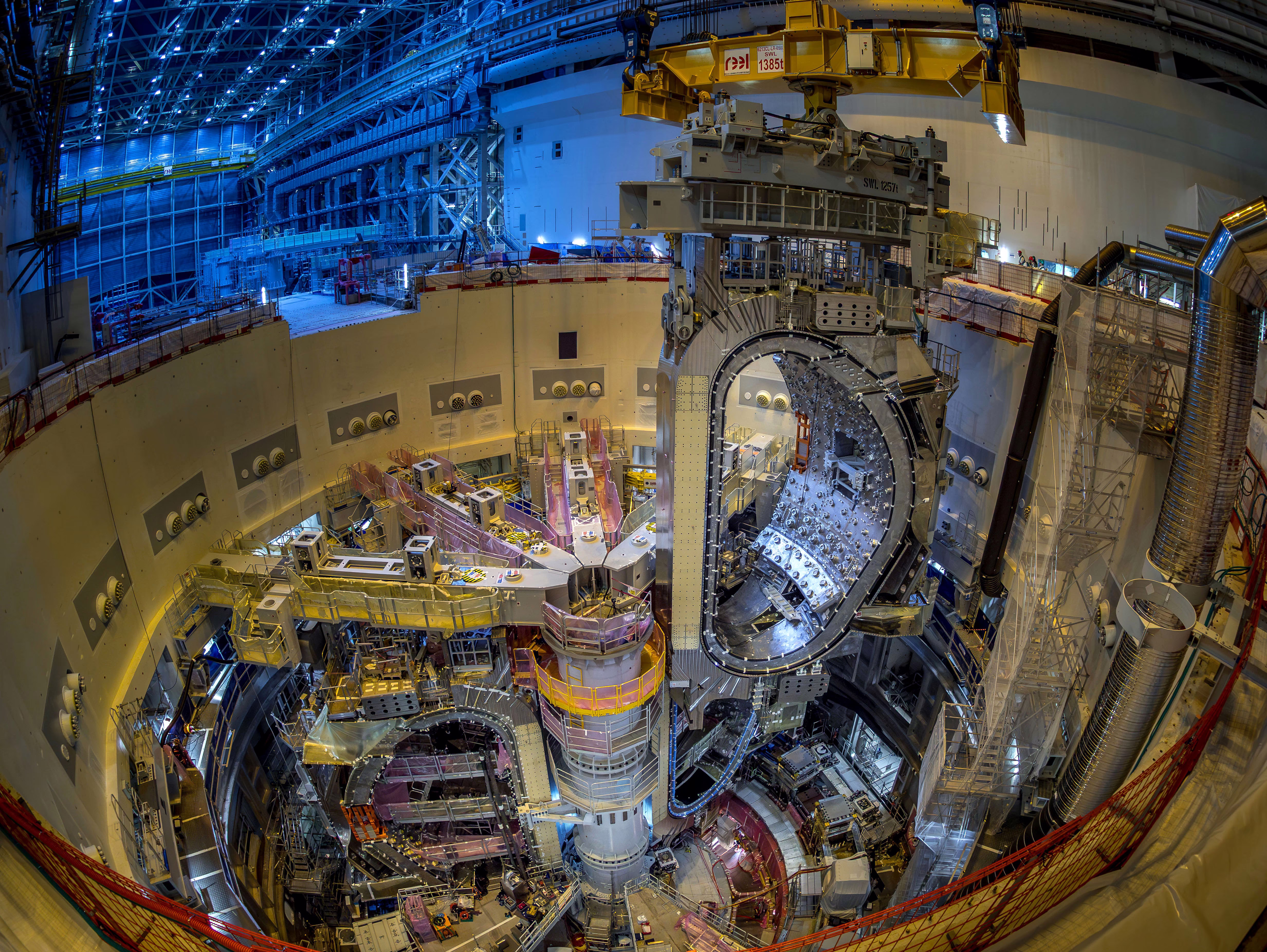A gantry crane lowers a large D-shaped component of a fusion reactor into place in a circular pit beside other components.