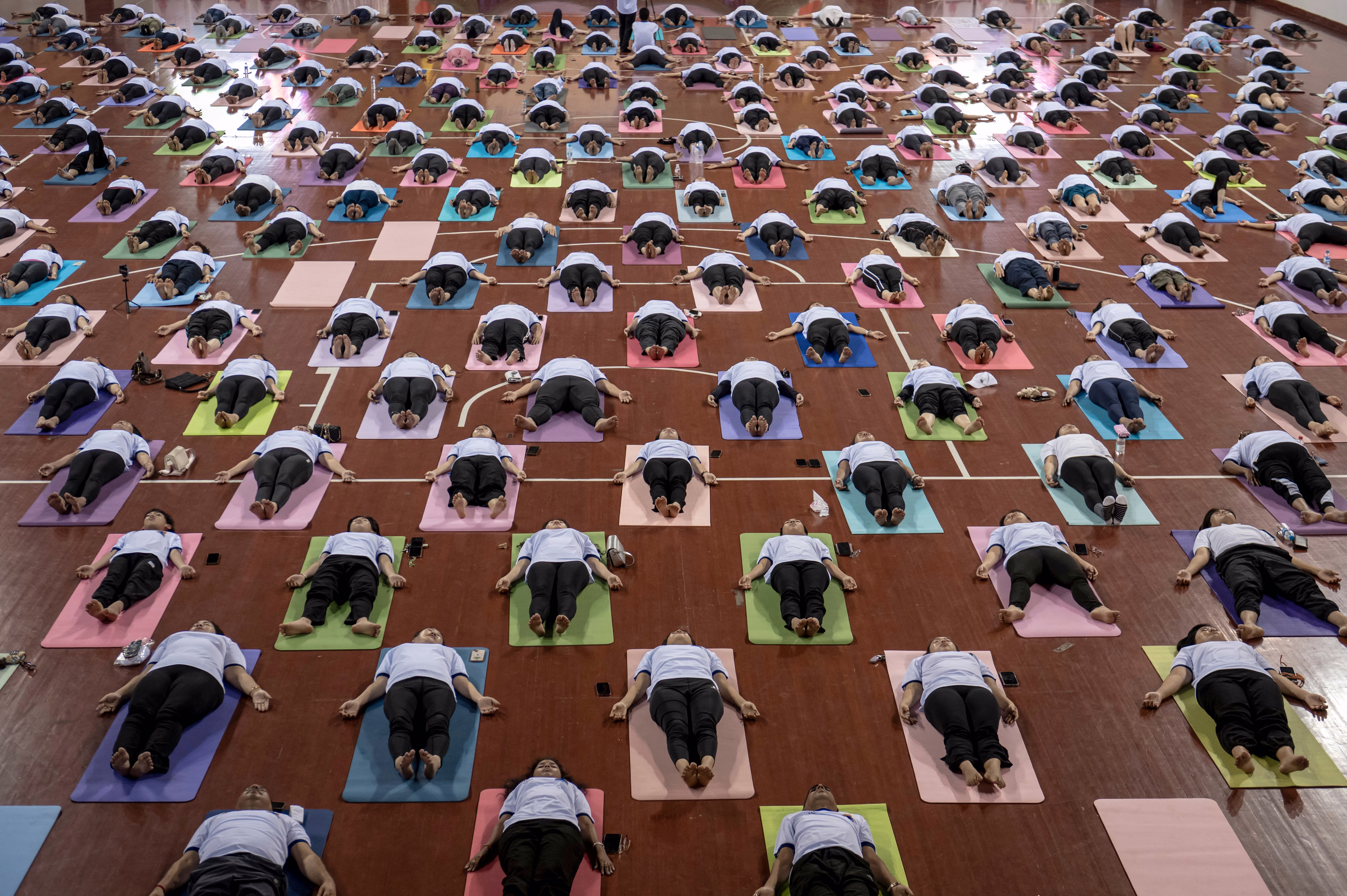 Dozens of people lie on their backs on yoga mats arranged on a large wooden floor in a grid pattern.