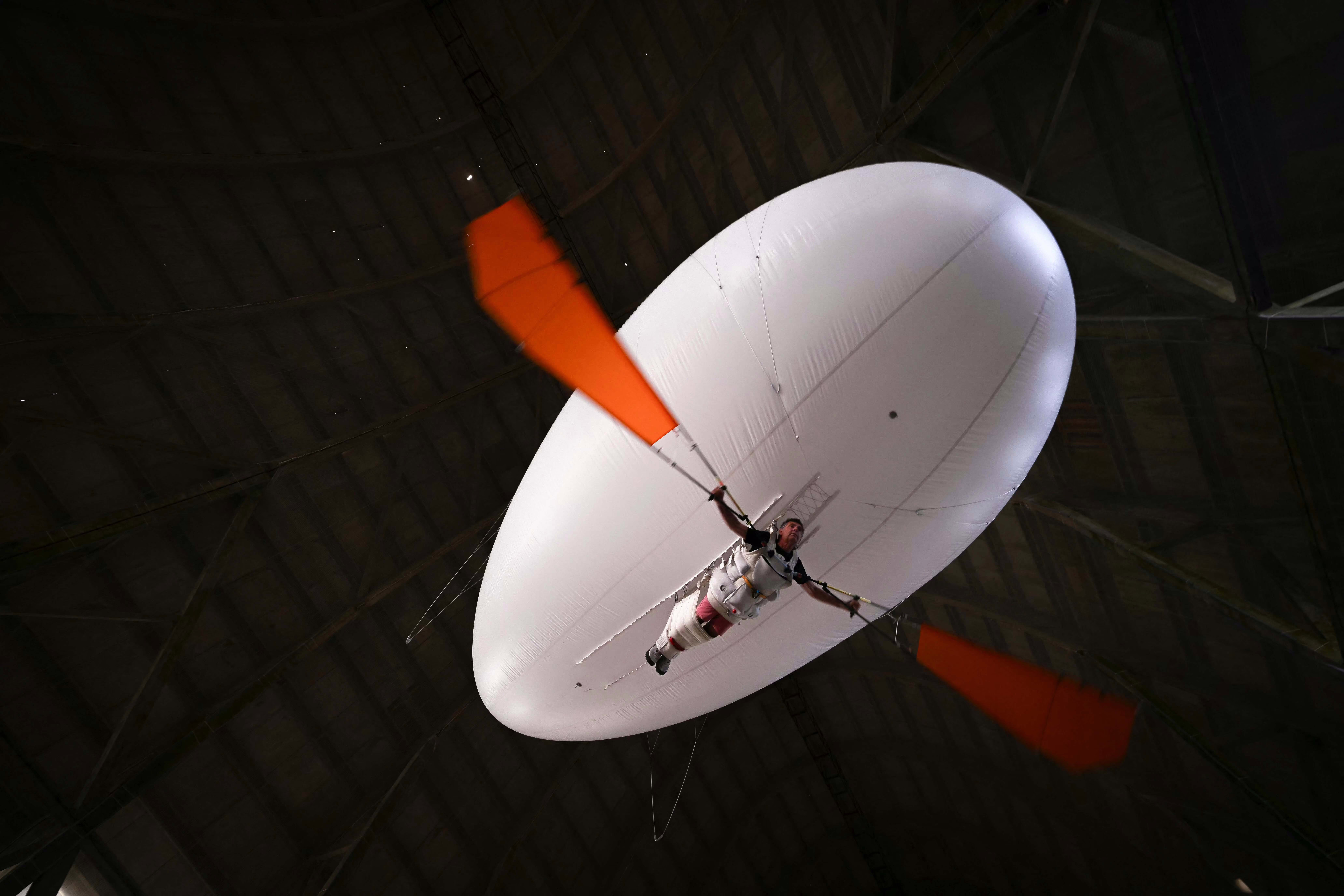A man hangs beneath a large balloon, strapped into a full-body harness, operating flaps with his arms, inside an airship hangar.