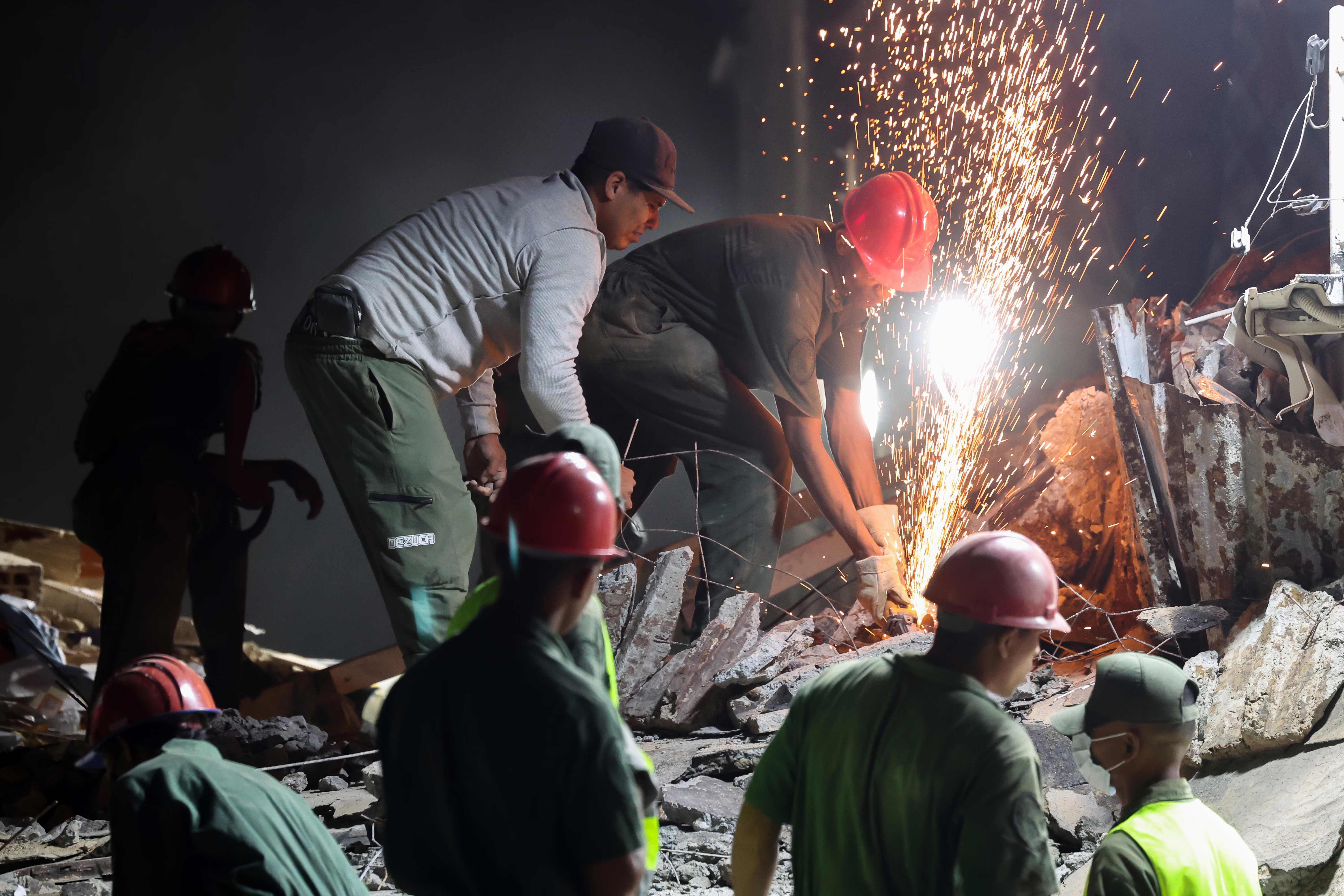 Sparks fly as a rescue worker uses a cutter to break apart metal reinforcing bars in a pile of rubble.