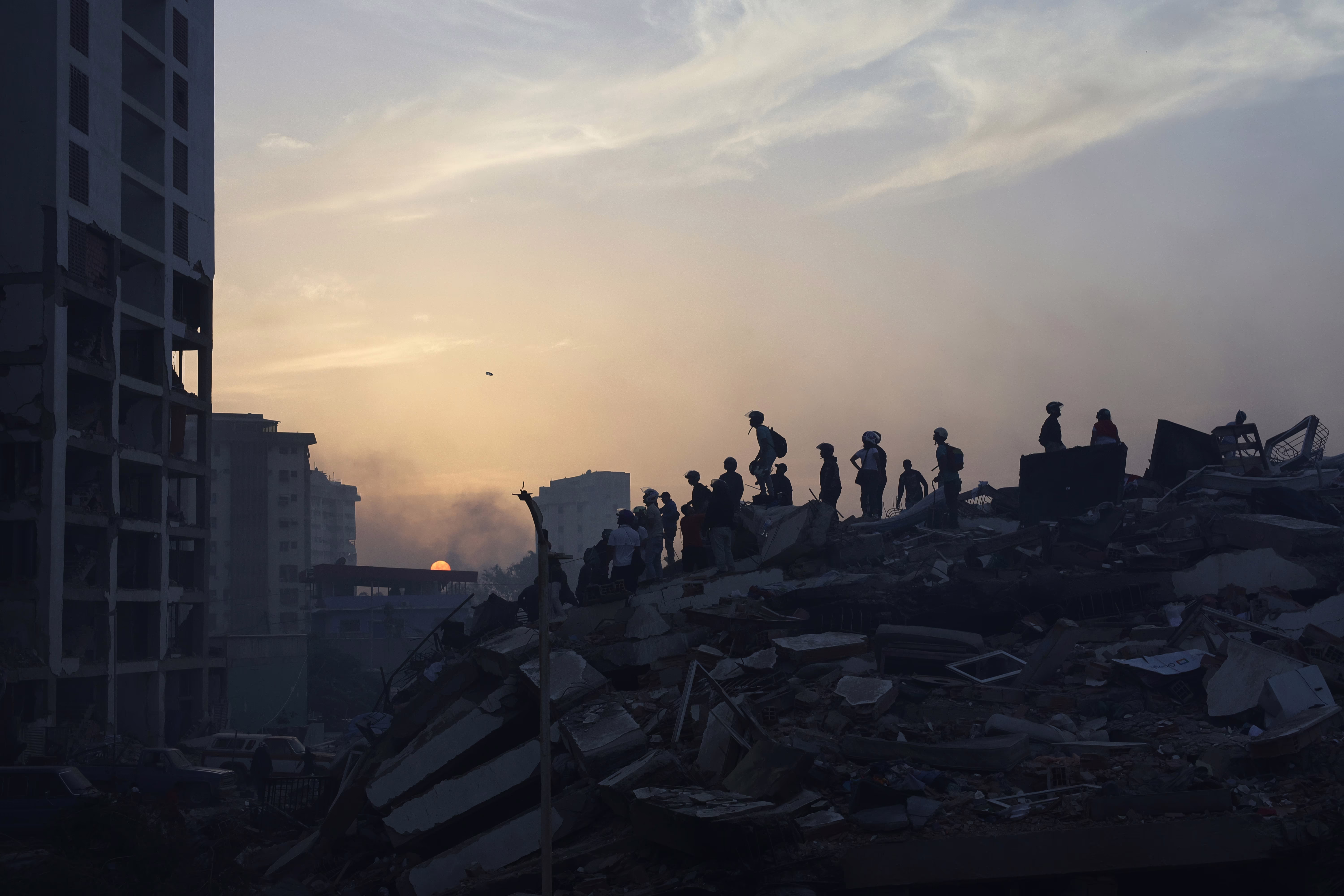 Rescue workers, seen in silhouette, stand atop a collapsed building.