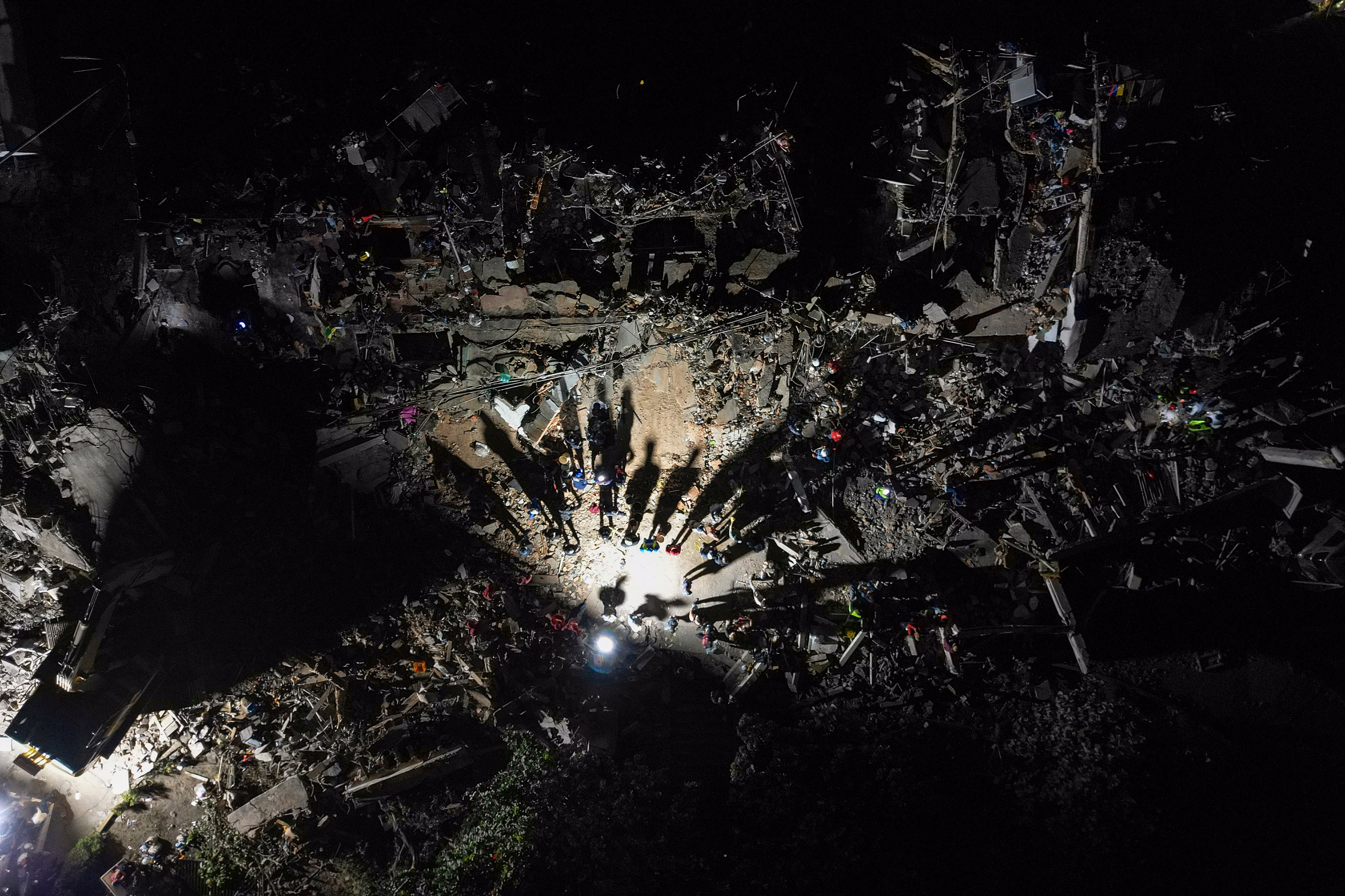 An aerial view of rescue workers amid destroyed buildings, seen at night, with long shadows cast by a work light.