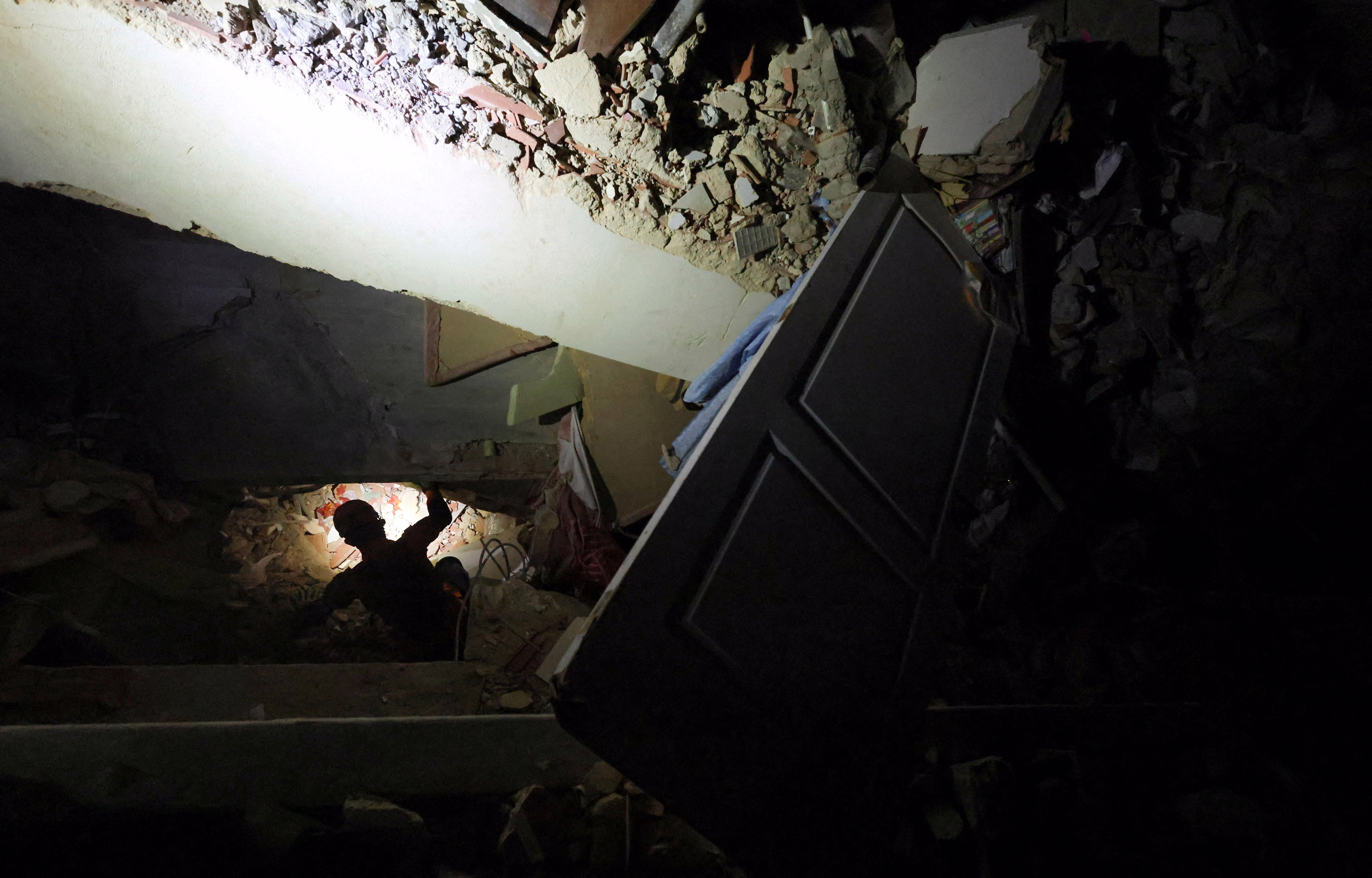 A rescue worker wearing a headlamp searches through the rubble of a large collapsed building in the dark.