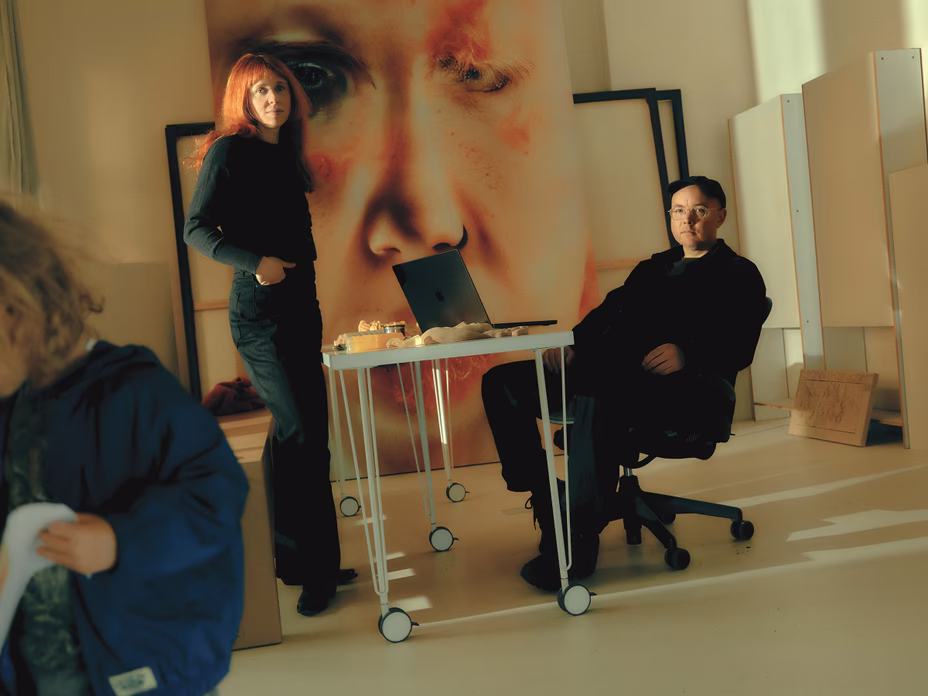 photo of woman standing and man sitting at table on wheels with large artwork leaning against wall behind and blur of toddler in foreground