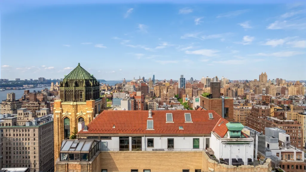 Rooftop view of a city with a distinctive Spanish-style villa atop a neo-Gothic church building, surrounded by other city buildings and a body of water in the distance.