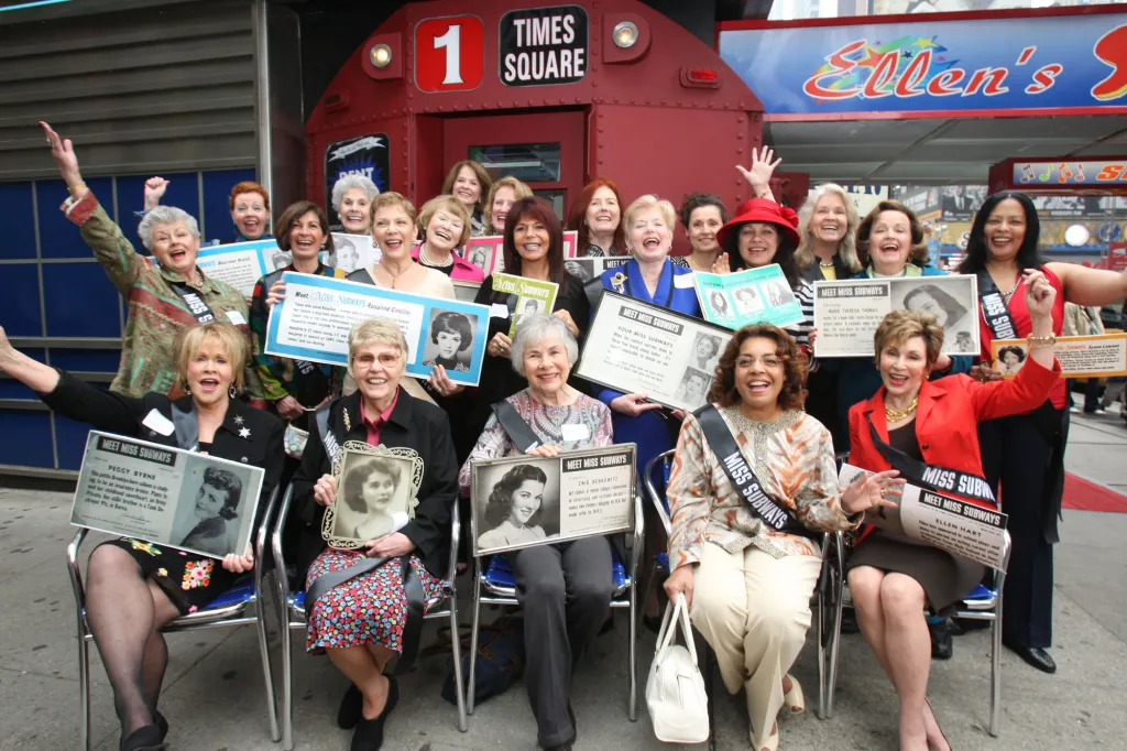 Former Miss Subways contest winners celebrate at a reunion in front of a red subway car display.