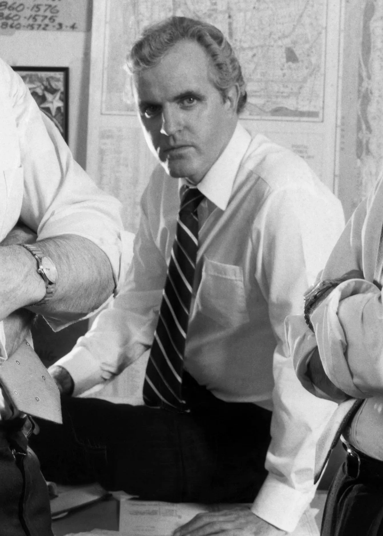 James Handy as a detective in a white shirt and striped tie, seated at a table.
