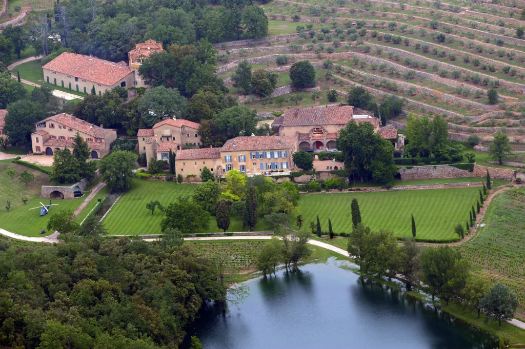 Aerial view of Chateau Miraval, a vineyard estate with buildings, lawns, terraced vineyards, a lake, and a helicopter.