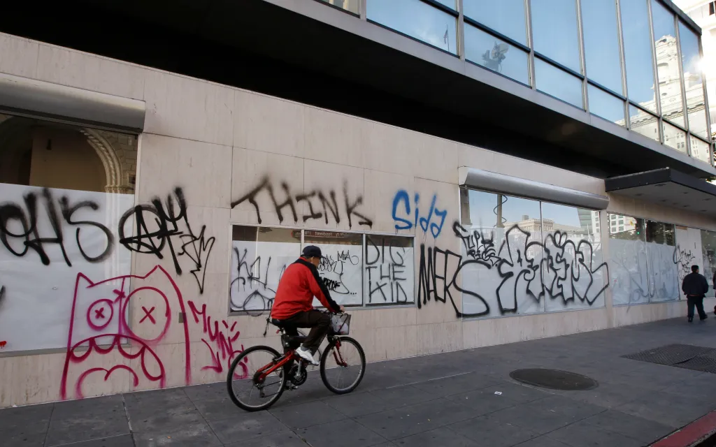 Graffiti covers a building facade in downtown Oakland, with a person riding a bicycle past the tagged windows.