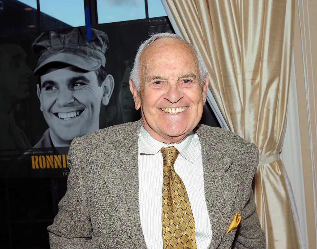 Ronnie Schell smiling, with a poster behind him of a younger version of himself in uniform.