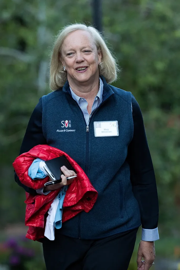 Meg Whitman, chief executive officer of Hewlett Packard, smiles while walking and holding a red jacket, smartphone, and pen.