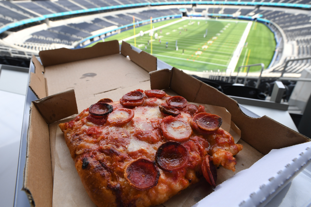 Marinara pizza with pepperoni and cheese in a cardboard box, with a view of a stadium field in the background.