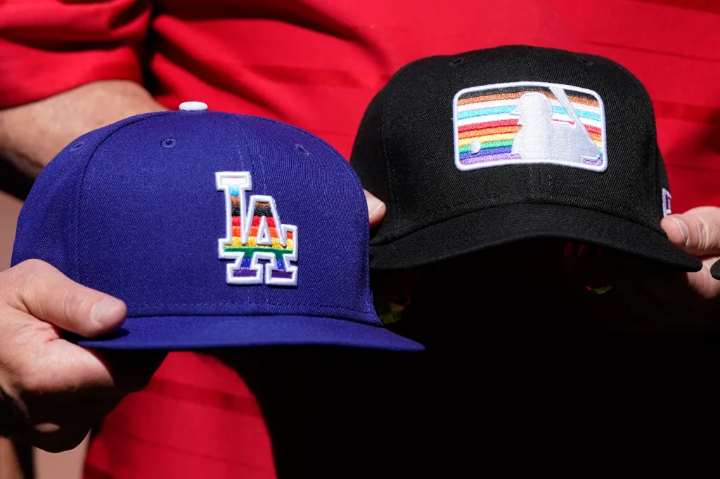 Two Pride-themed baseball hats, a blue Los Angeles Dodgers hat and a black Major League Baseball hat, are displayed.