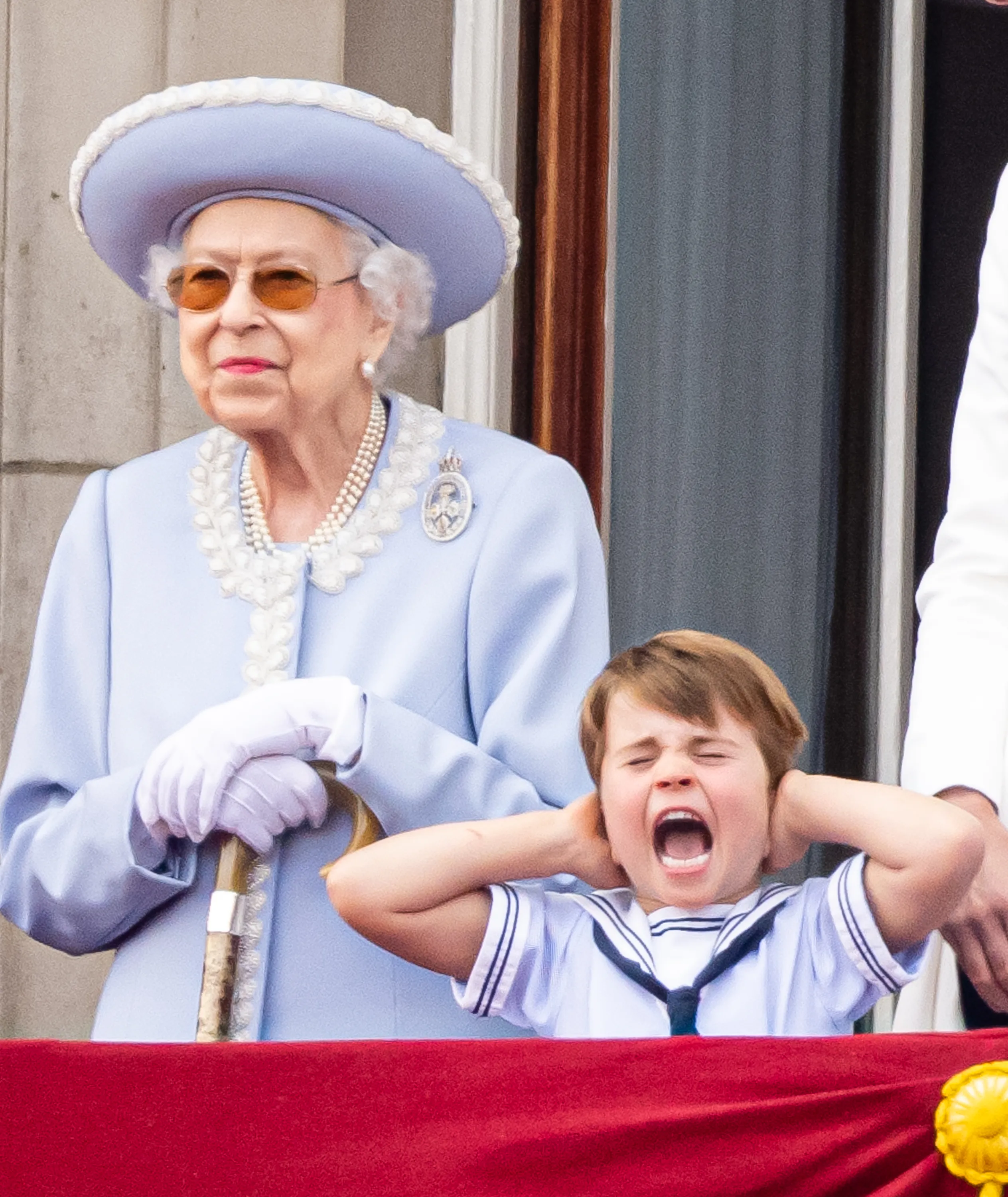 Queen Elizabeth II in a light blue outfit with a cane standing next to a boy in a sailor suit with his hands over his ears and mouth open, shouting.