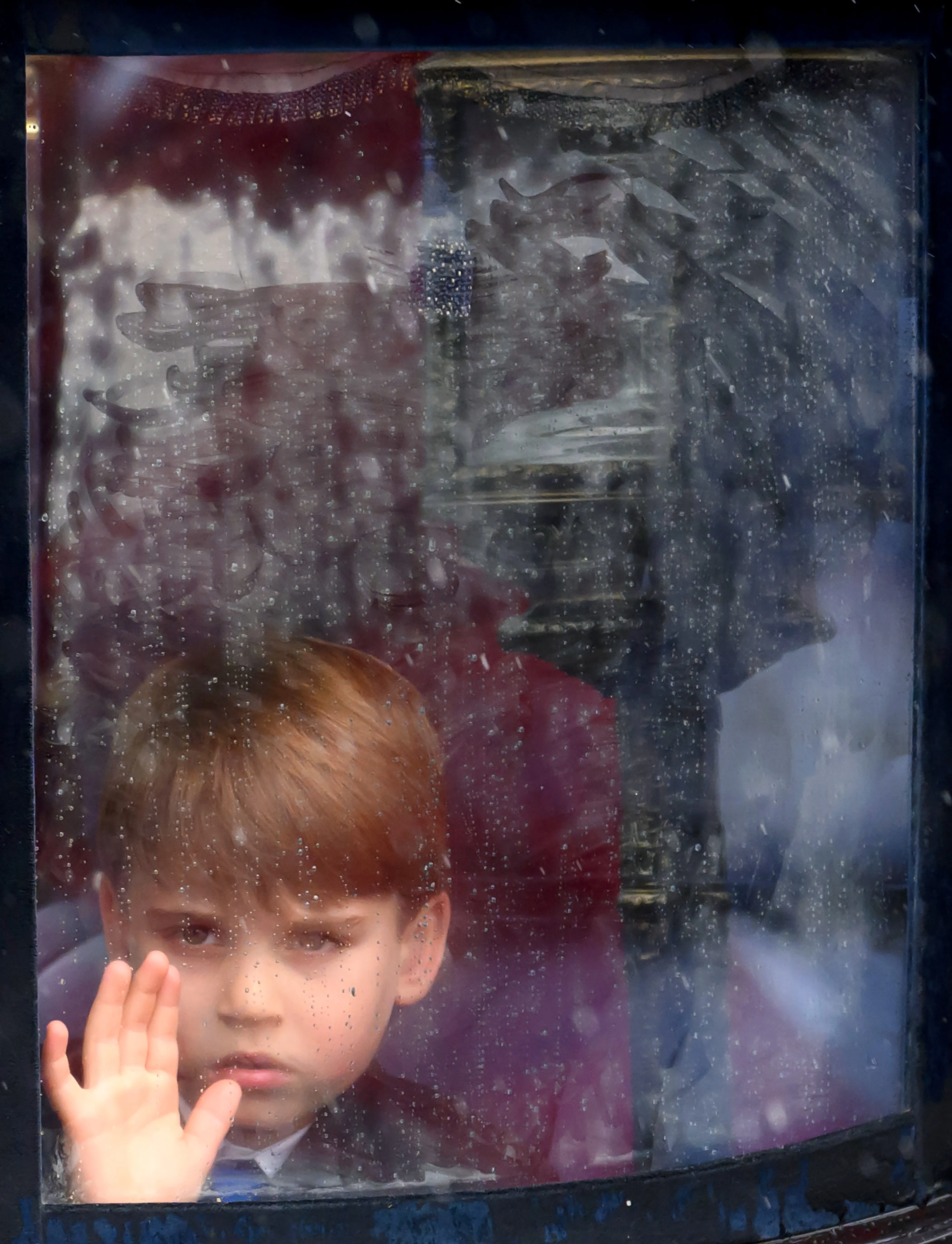 Prince Louis of Wales looks through a wet window, with his hand pressed against the glass.