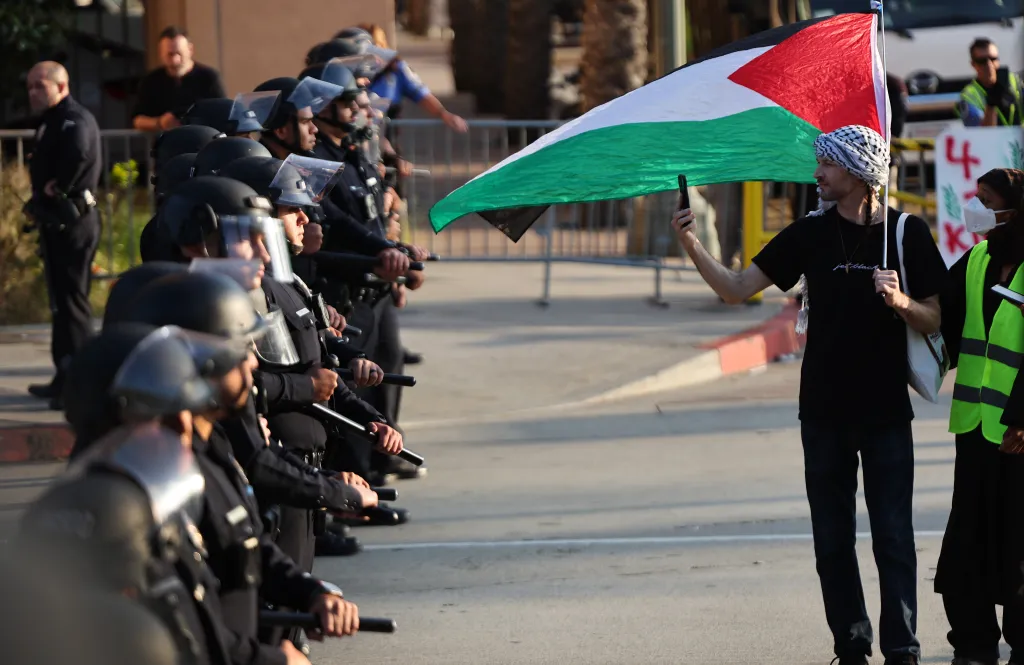 A pro-Palestinian protestor holding a Palestinian flag walks past a line of LAPD officers.