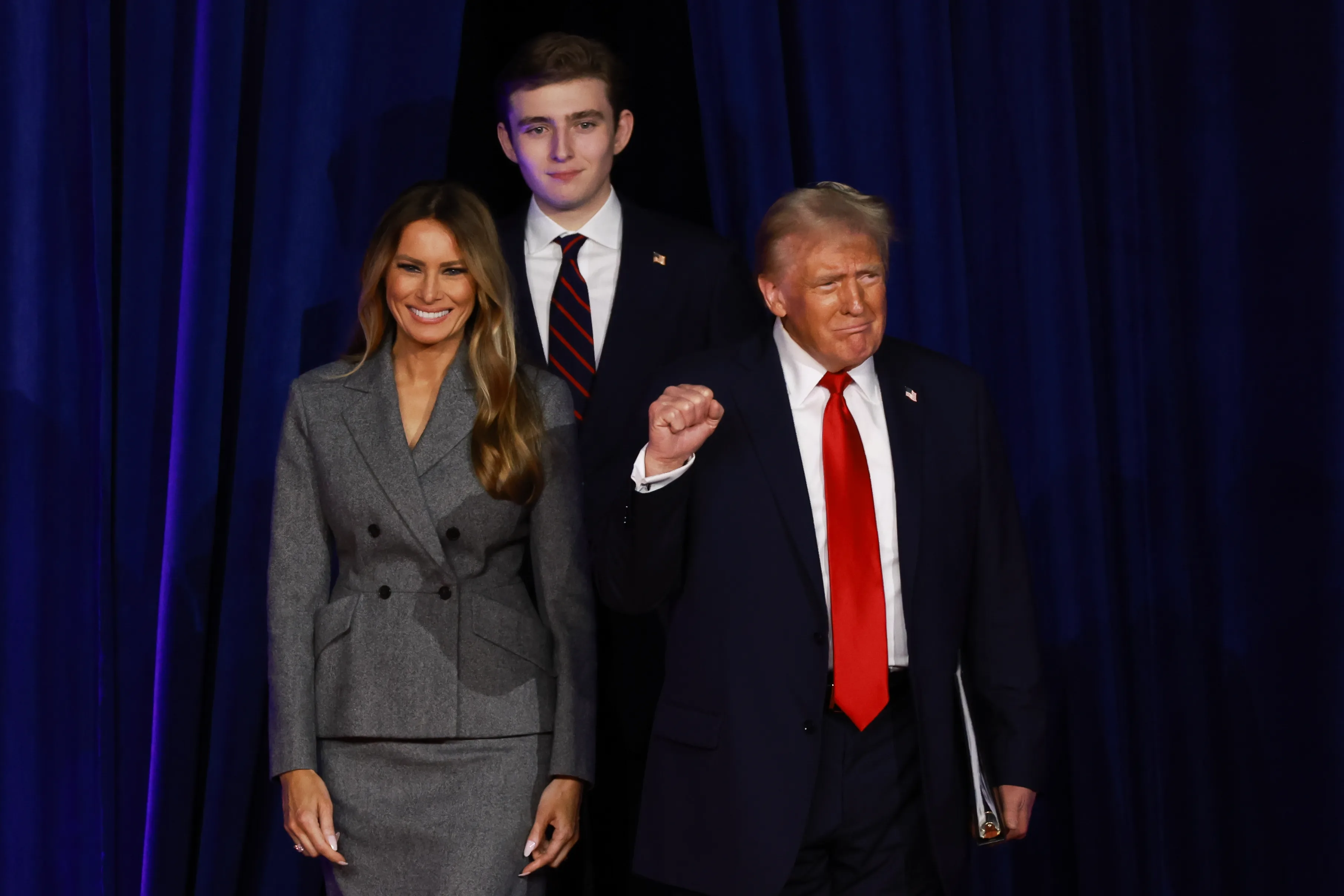 Donald, Melania, and Barron Trump walk across a stage with blue curtains in the background.