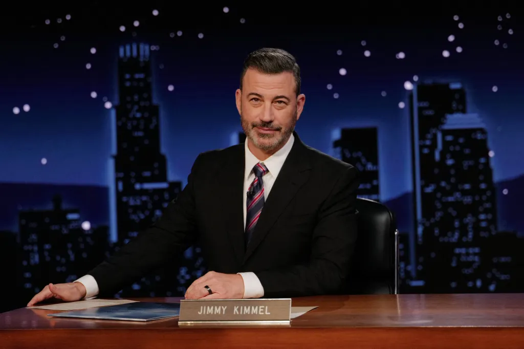 Jimmy Kimmel seated at a desk, smiling, with a city skyline background.