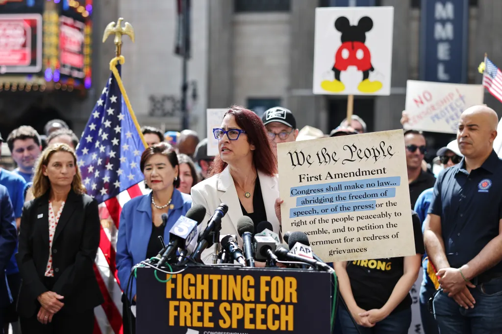 U.S. Rep. Laura Friedman speaks at a podium with microphones, holding a sign displaying the First Amendment, during a protest against the suspension of the 