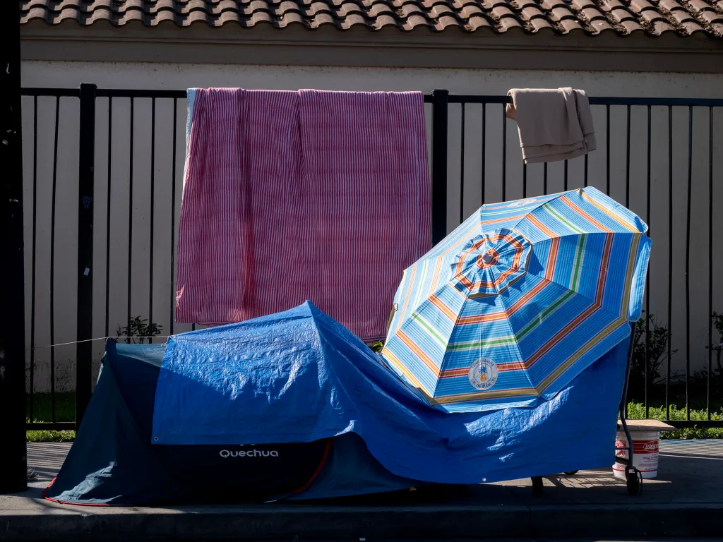 Homeless encampment in Santa Monica with a tent, blue tarp, and striped umbrella by a fence.