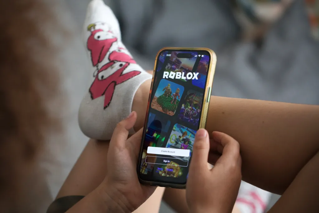 A child holds a smartphone displaying the Roblox app login page. 