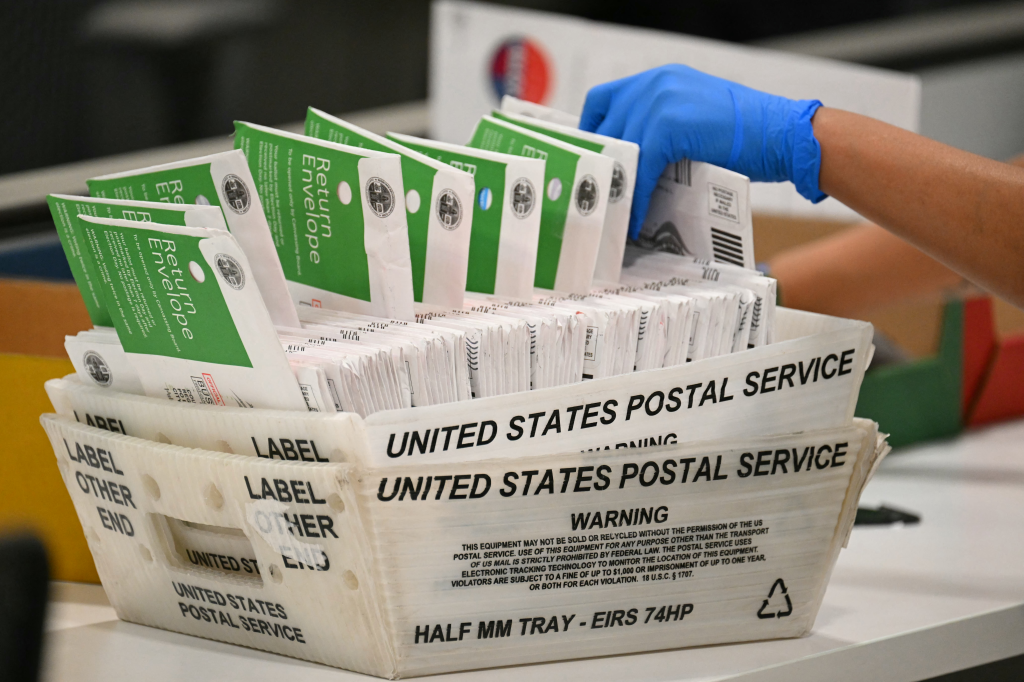 An election worker wearing a blue glove processes mail-in ballots in a United States Postal Service bin.