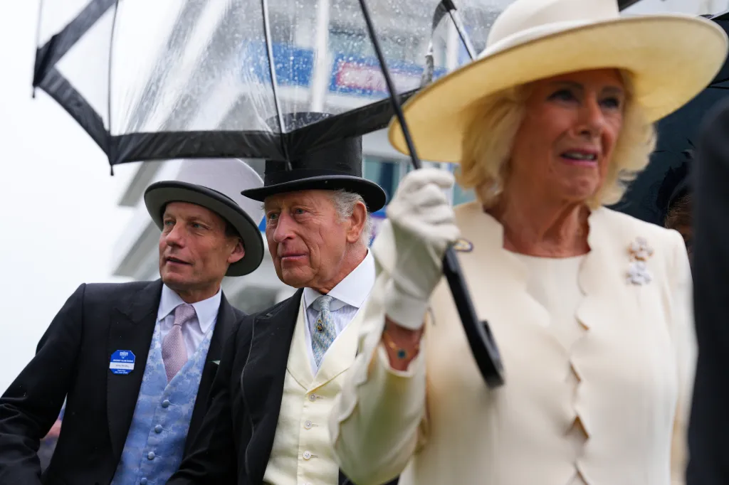 King Charles III and Queen Camilla attend Derby Day at Epsom Downs Racecourse, with Andy Hornby alongside them under umbrellas.