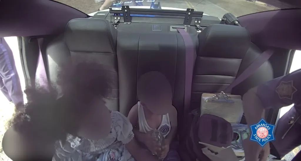 View from inside a police vehicle shows two children and an adult police officer in the back seat, with a clipboard and Arkansas State Police logo visible.