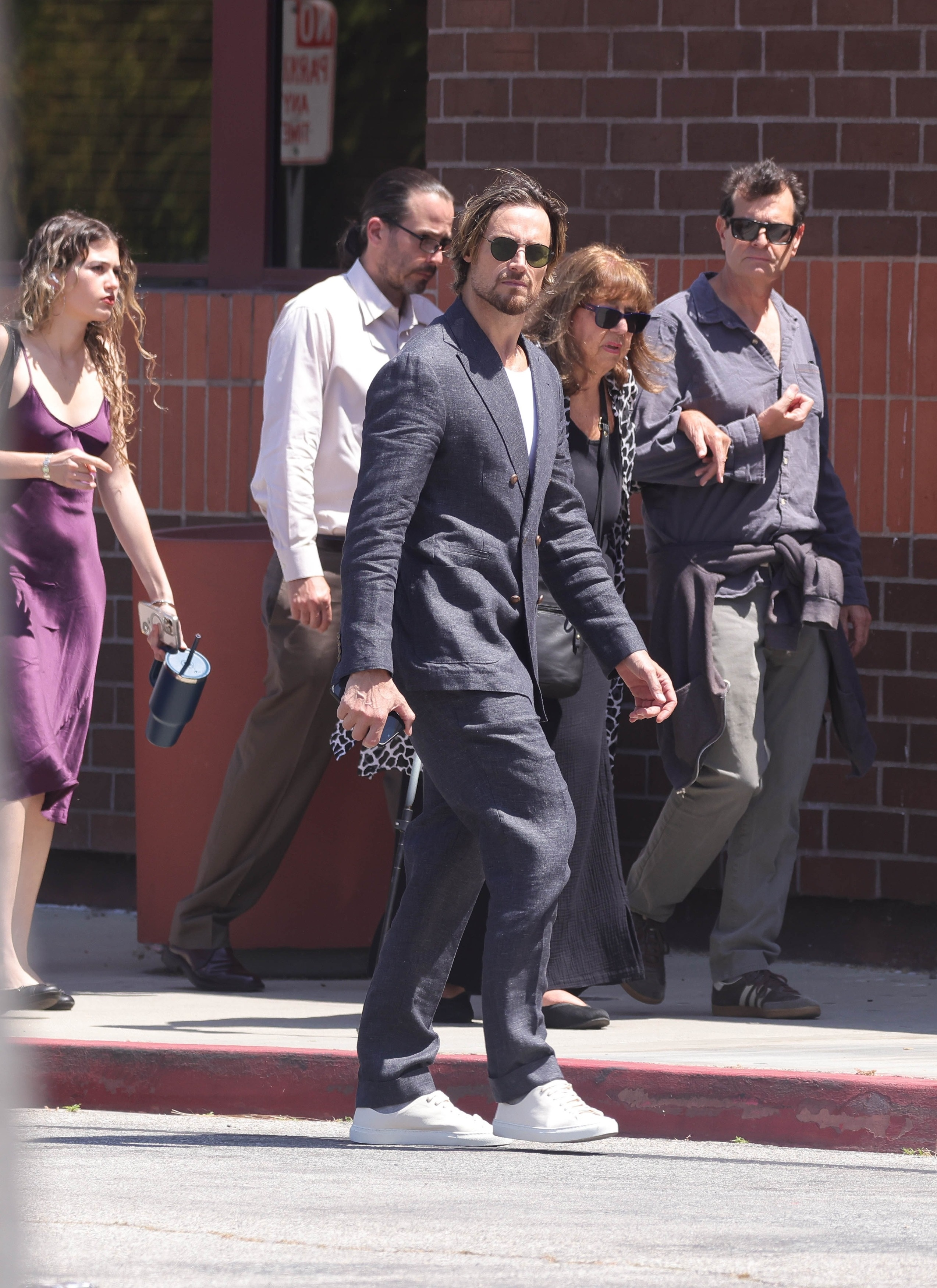 Gabriel Aubry in a grey suit, white sneakers, and sunglasses, walking with a group of people.